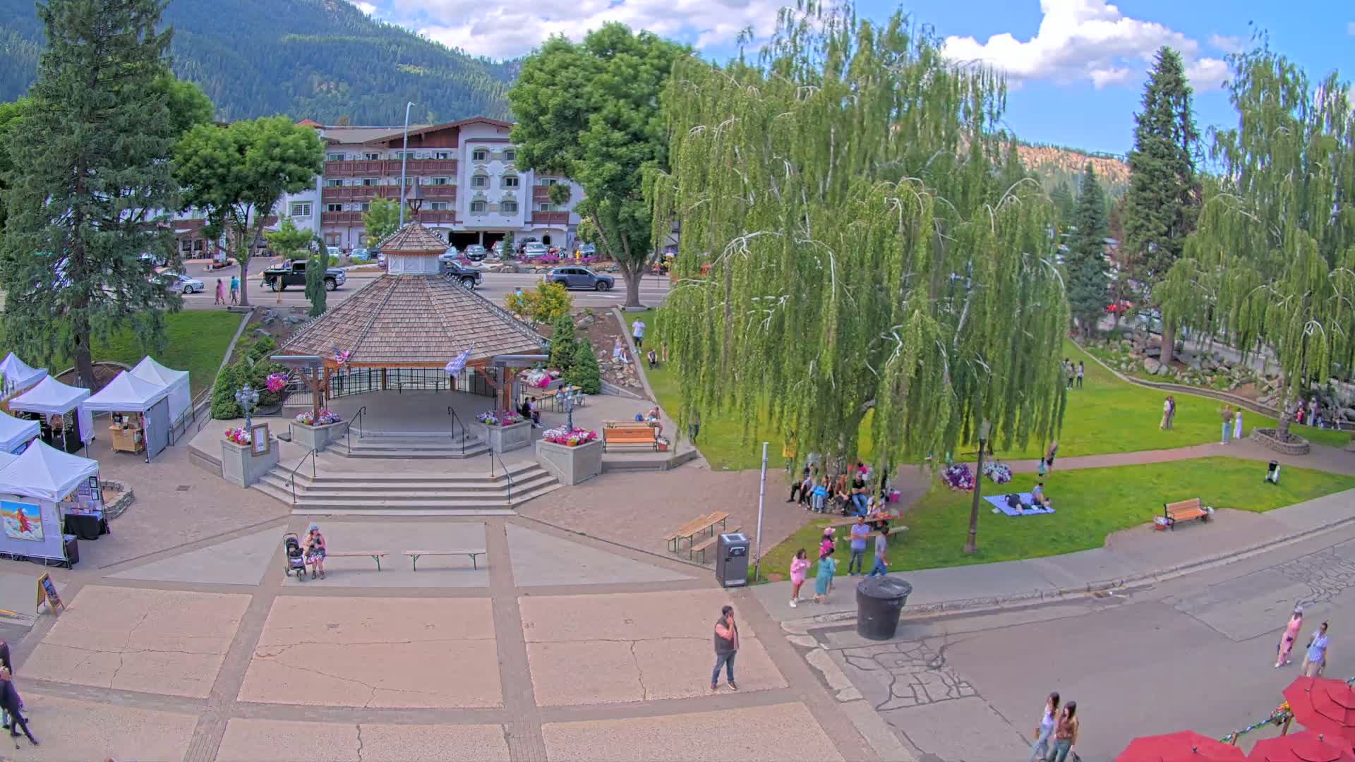 A town square with a gazebo, surrounded by green grass, trees, and a few people, under a clear sunny sky, features a small art market and a hotel in the background.