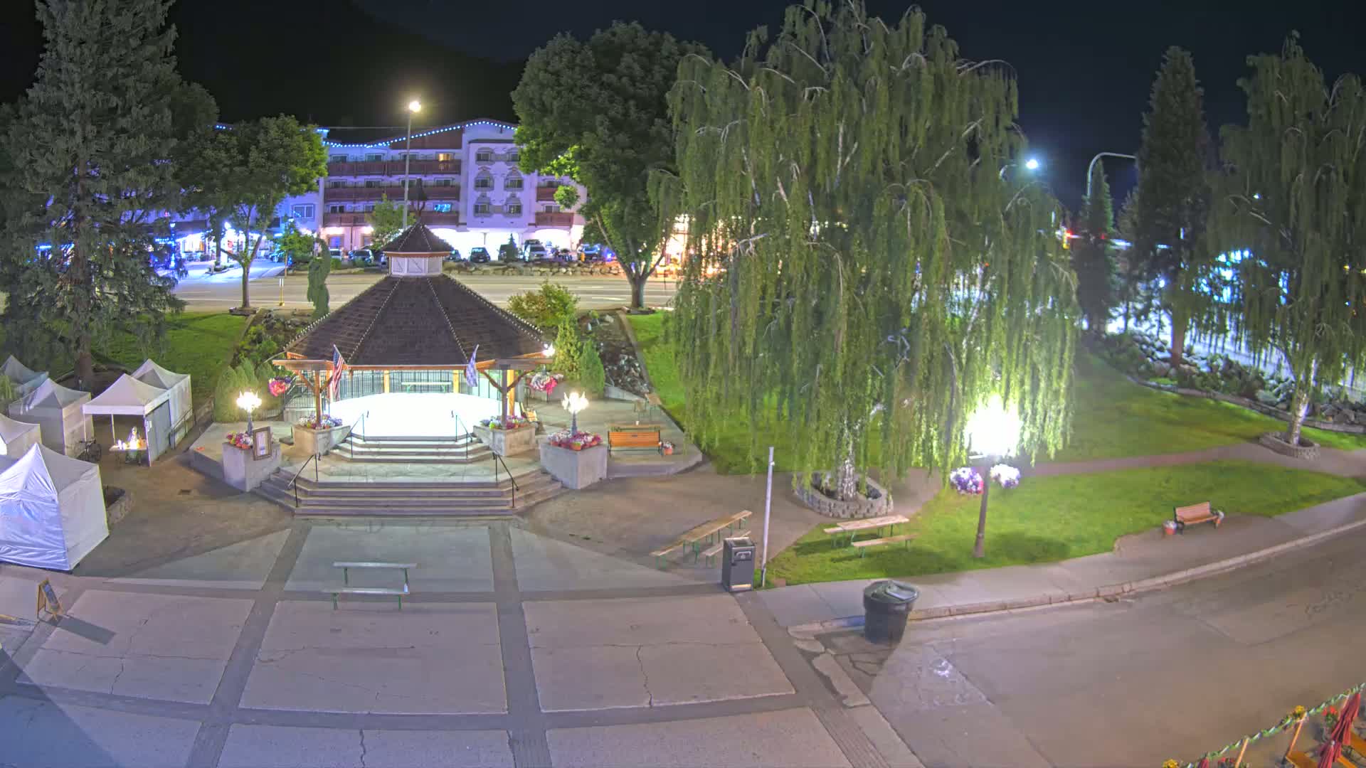 A nighttime view of a town square with a gazebo, several small tents, and a few benches, illuminated by streetlights, next to a multi-story building.