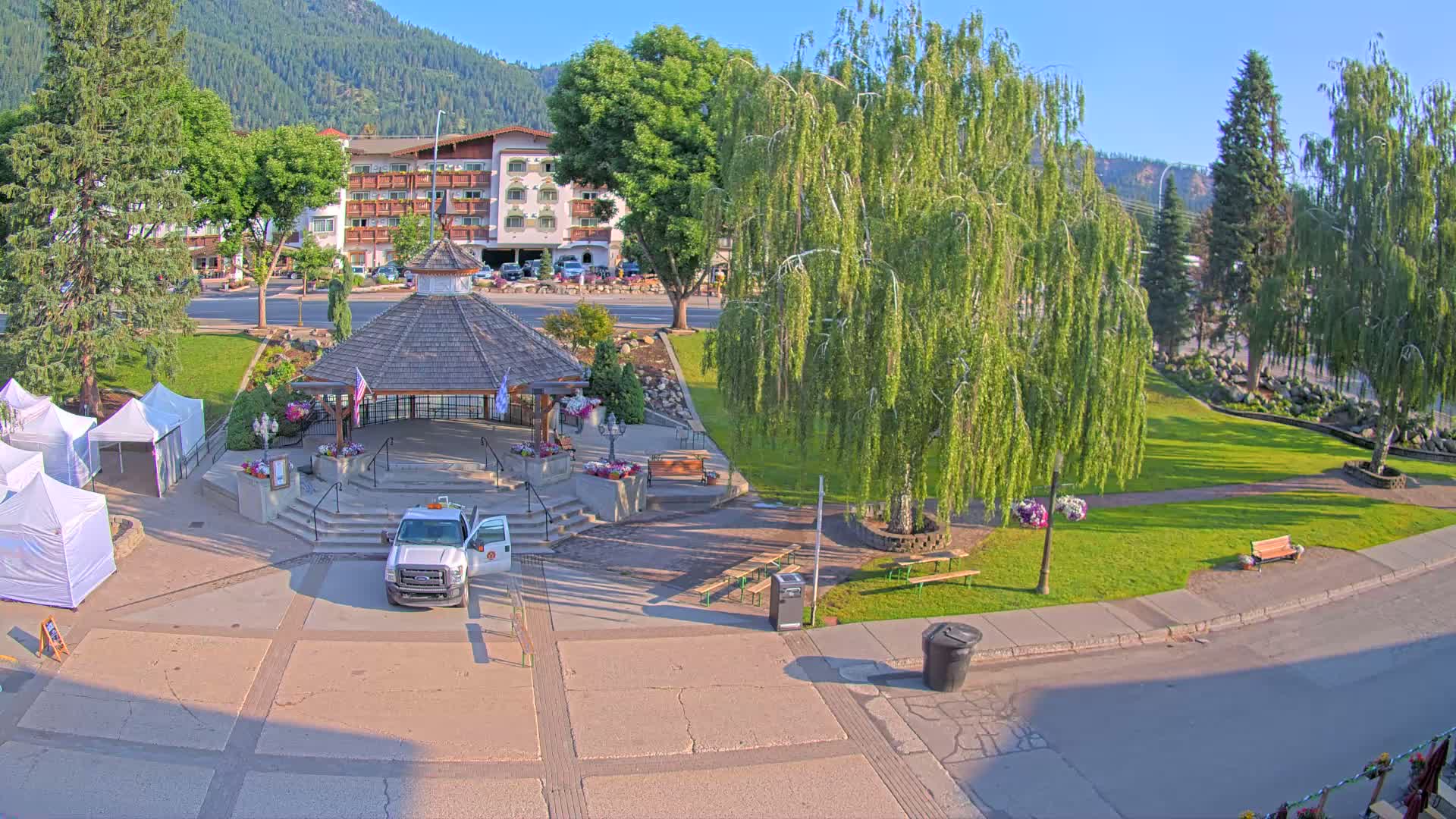 A white truck is parked near a gazebo in a park with green lawns, leafy trees, and a building in the background, all under a sunny sky.