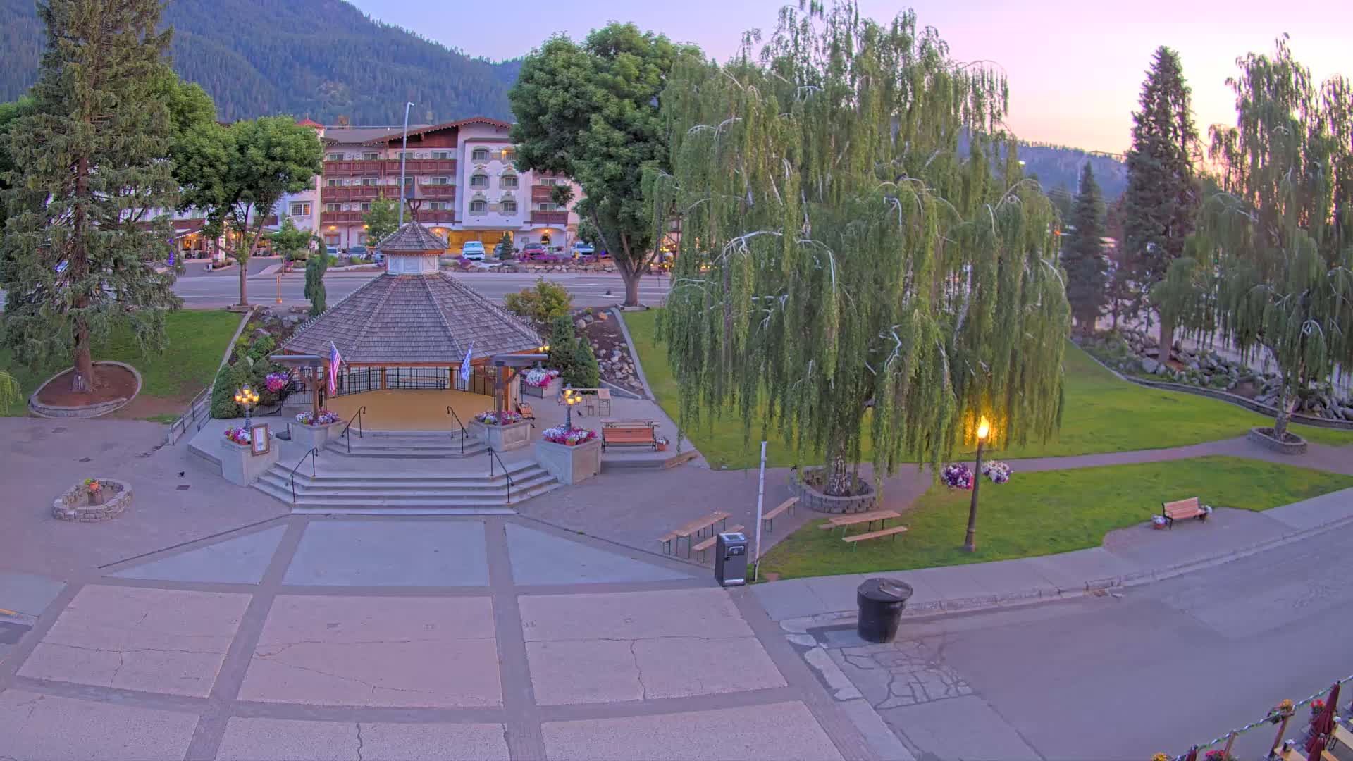 A gazebo with a shingled roof sits in a park at dusk, next to a weeping willow tree and a multi-story hotel, with mountains visible in the background.