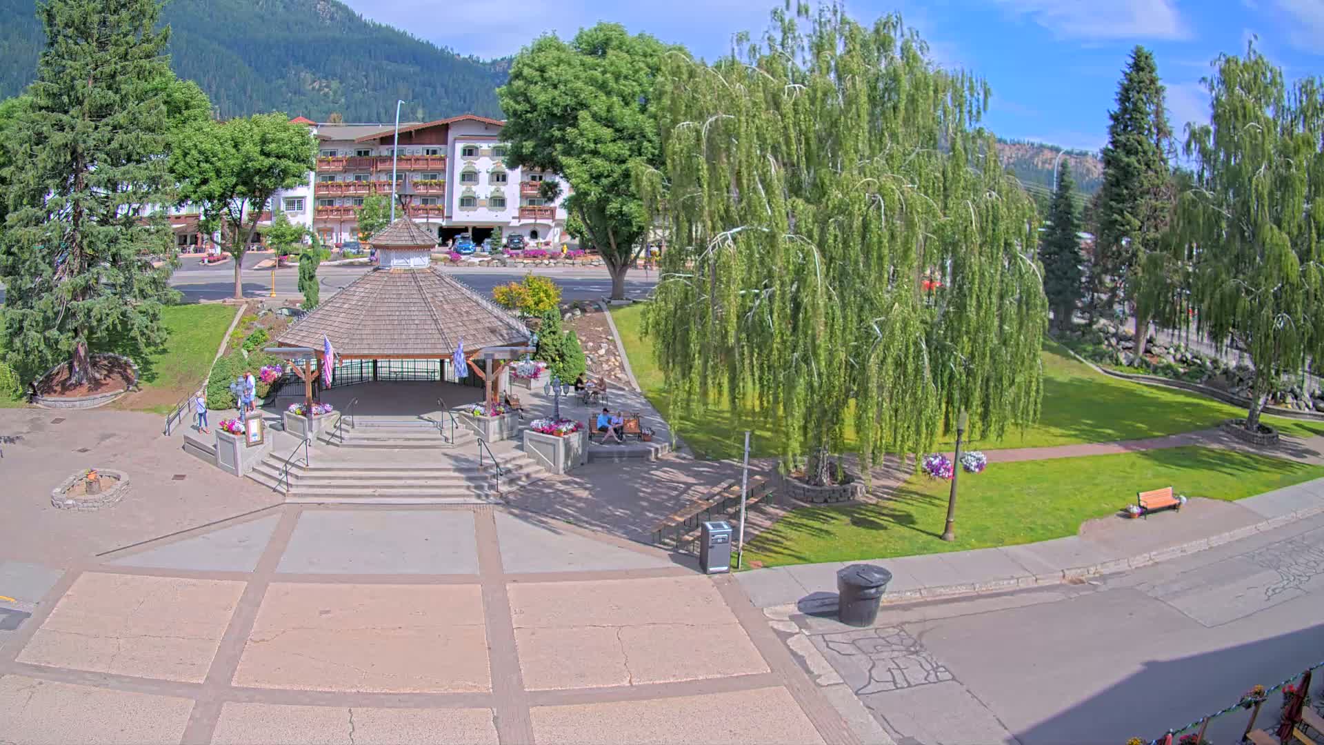 A park with a gazebo and several people sitting on benches is situated in front of a multi-story building, all under a sunny sky.