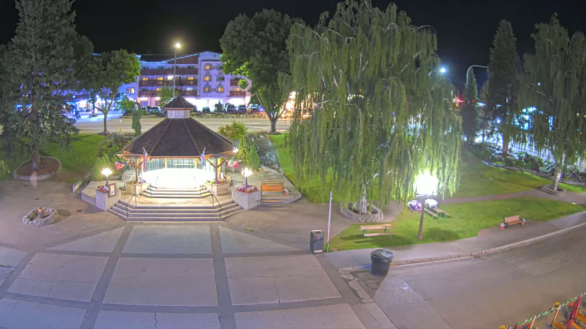 A nighttime aerial view shows a gazebo in a park, illuminated by lights, next to a roadway and a multi-story building.