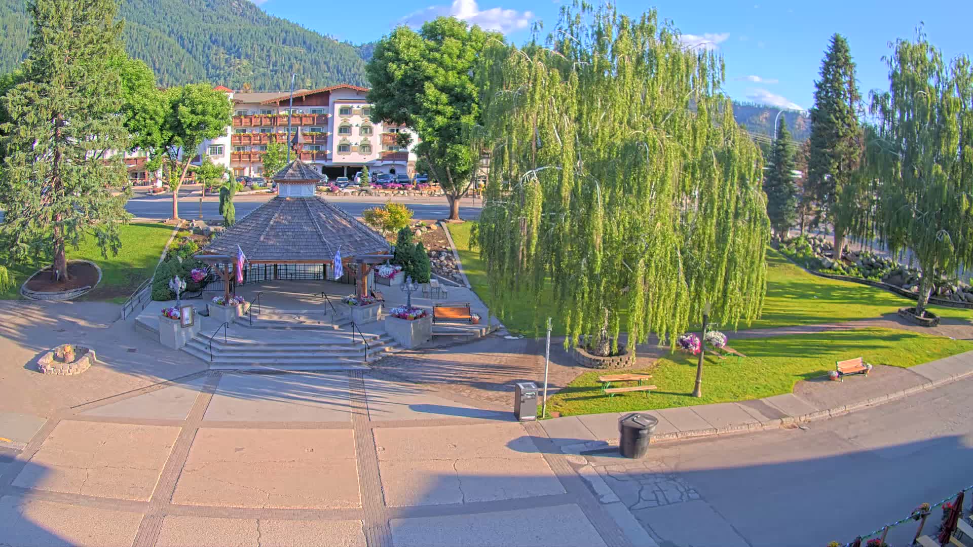 A paved town square with a gazebo, benches, and several trees, situated in front of a hotel, on a sunny day with clear skies and mountains in the background.