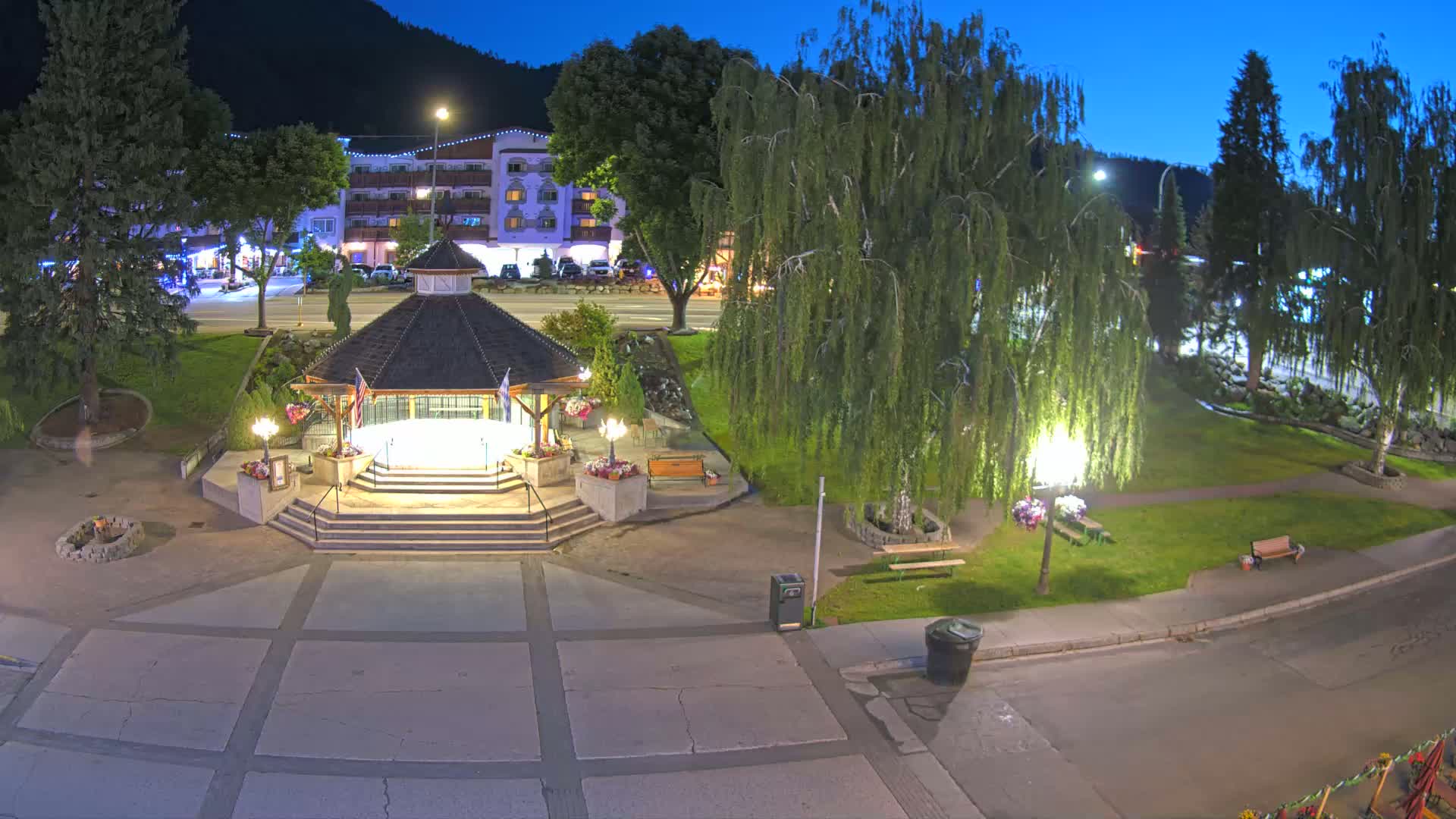 A nighttime view of a town square with a gazebo, trees, benches, and a building in the background under a twilight sky.