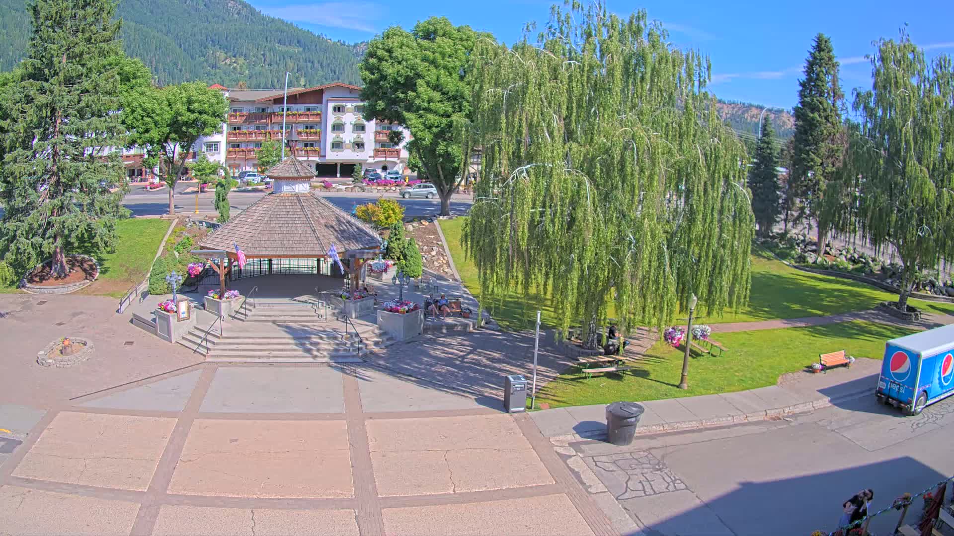 A park with a gazebo, trees, and benches sits in front of a hotel on a sunny day with mountains in the background.