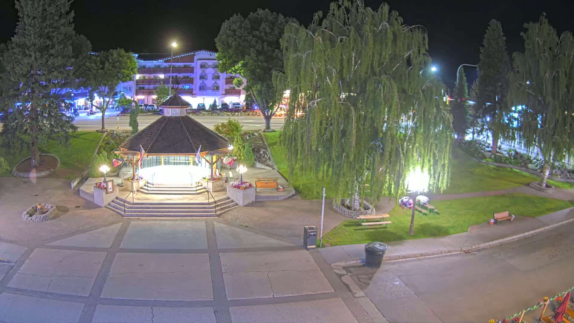 A nighttime view of a town square with a gazebo, surrounded by trees and a building, under clear skies.