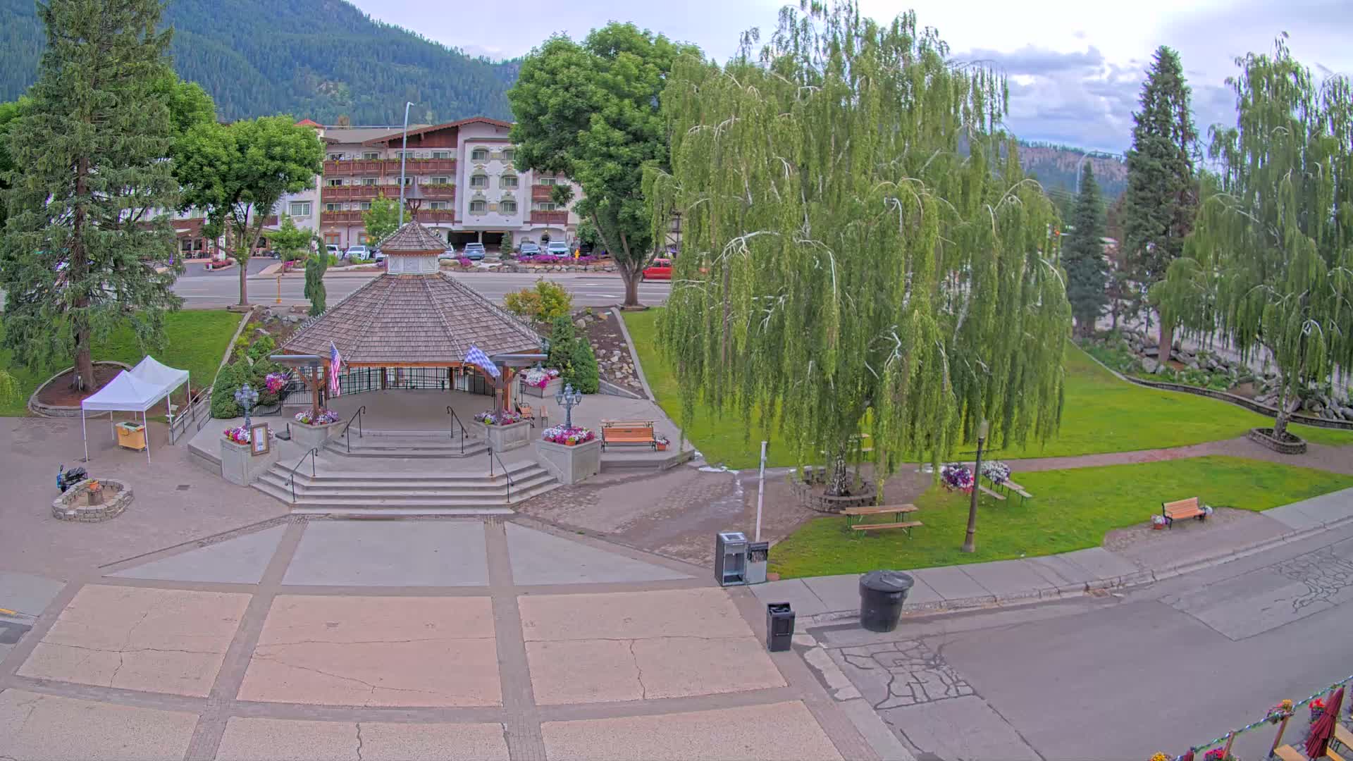 A gazebo with stairs sits in a town square, surrounded by green grass, trees, and a multi-story building, under a partly cloudy sky.