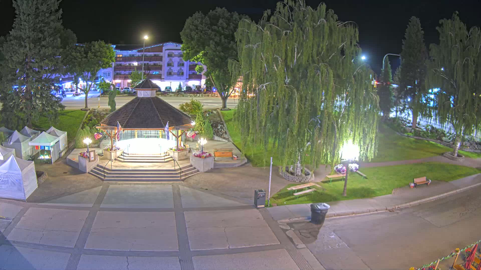 A nighttime view of a town square with a gazebo, several small tents, a park with trees, and a street with buildings in the background.