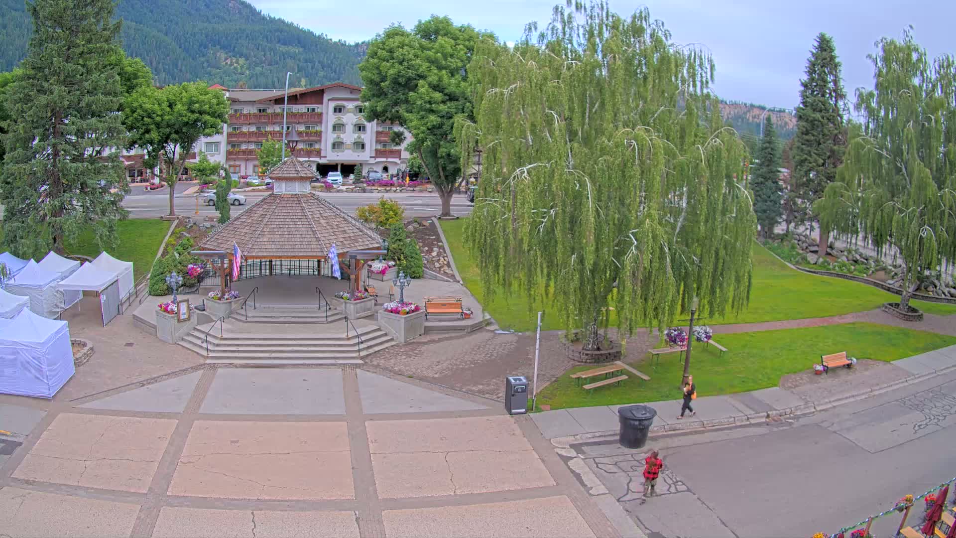 A town square with a gazebo, park benches, a few people walking, and white tents set up on a partly cloudy day in front of a hotel and mountains in the background.
