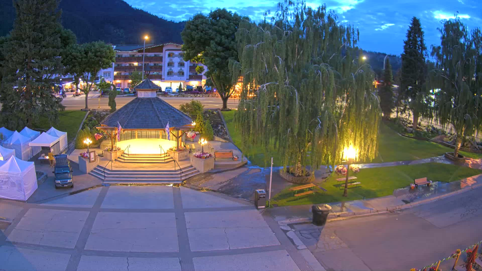 A town square at dusk features a gazebo, several park benches, and some small white tents under a partly cloudy sky.