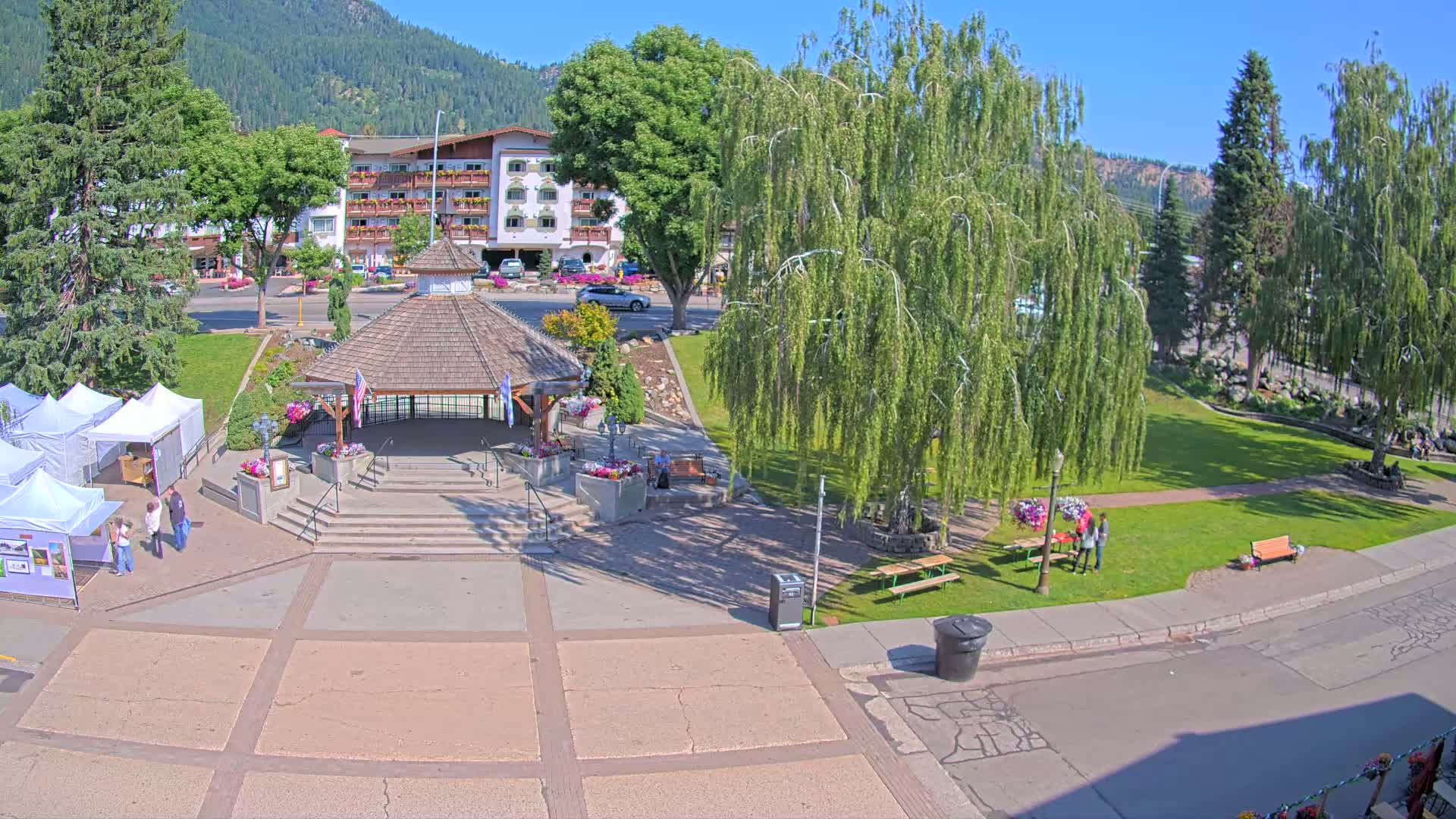 A town square with a gazebo, several people, and white vendor tents under a sunny sky, surrounded by trees and a building.