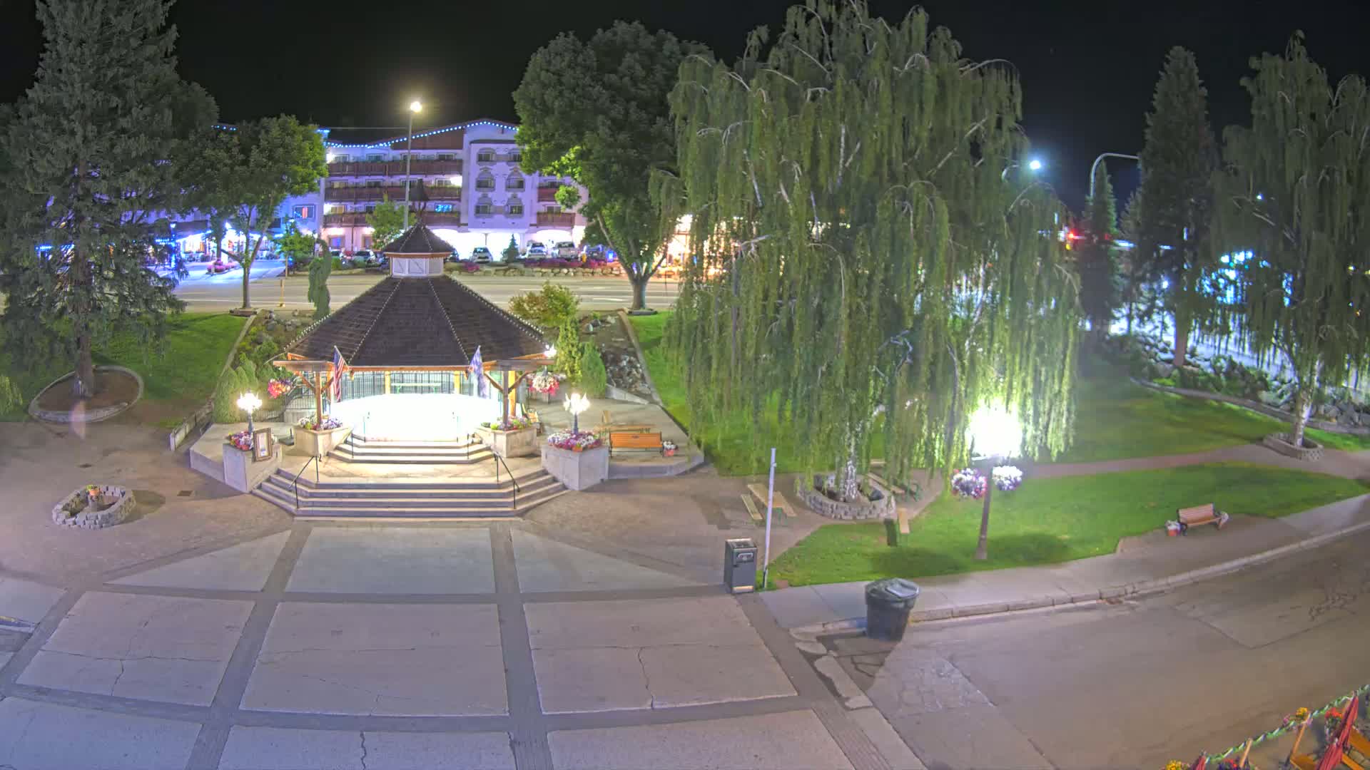 A nighttime aerial view shows a park with a gazebo, benches, trees, and a paved area, next to a street and buildings.