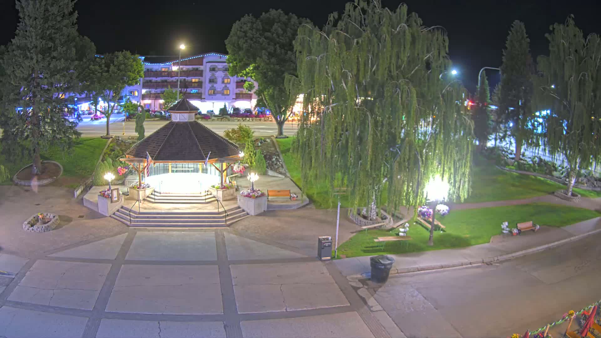 A gazebo sits in a park at night, illuminated by streetlights, with a street and buildings visible in the background.