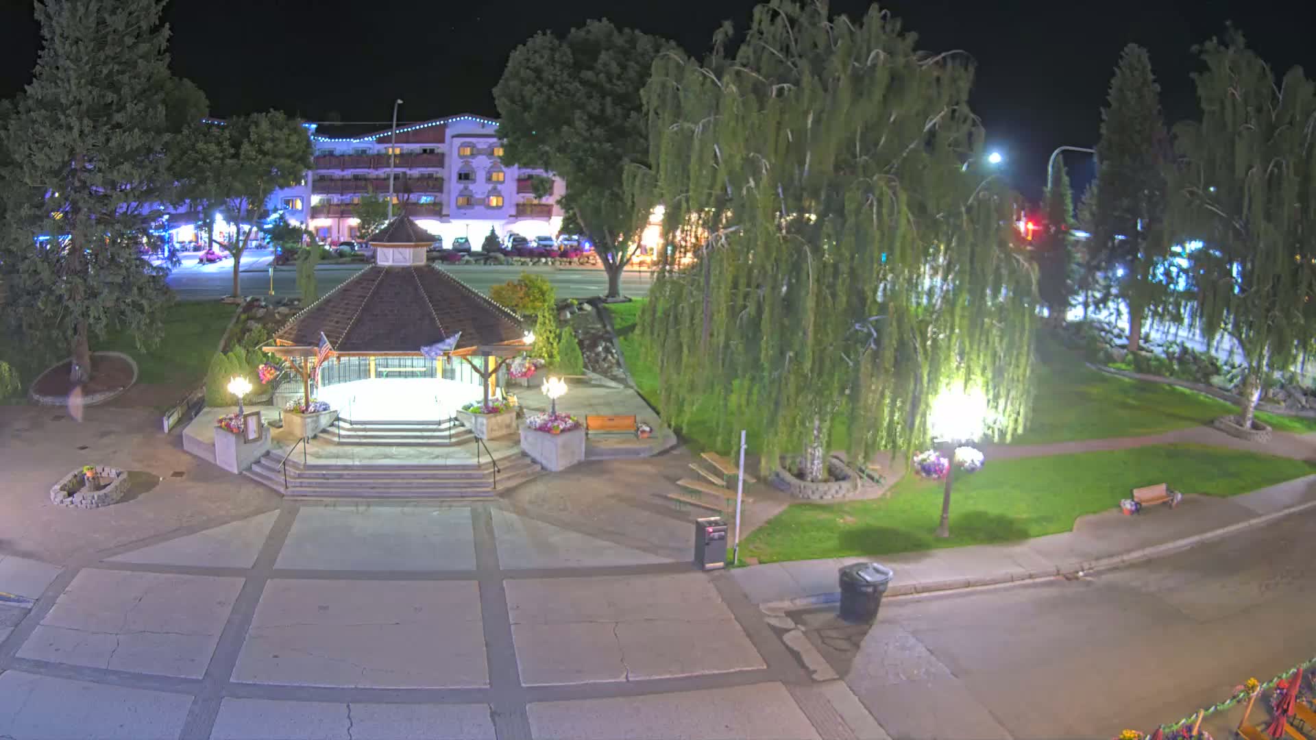 A nighttime view of a town square featuring a gazebo, landscaping, and a building with lights.