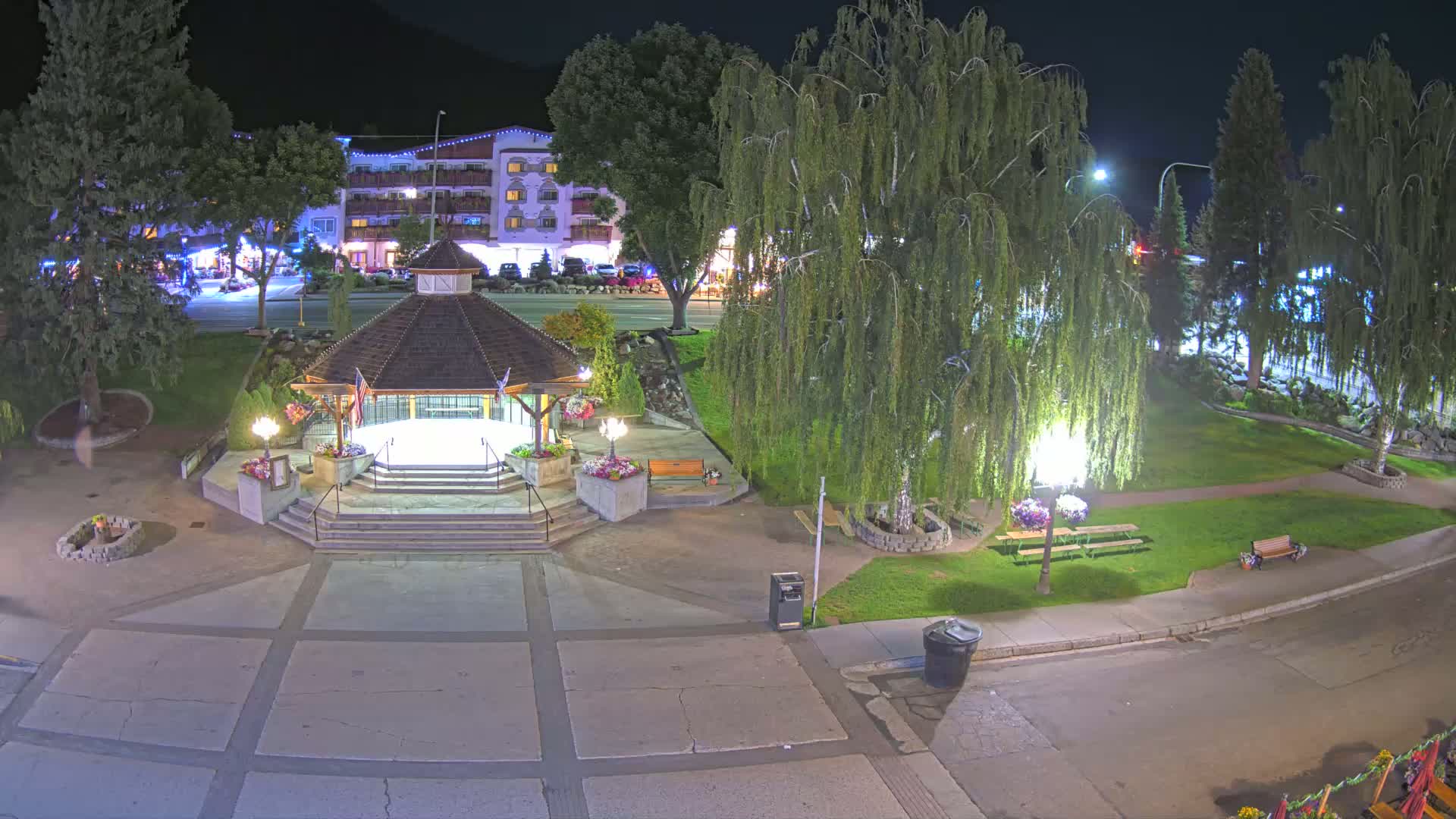 A nighttime view of a town square featuring a gazebo, landscaping, and streetlights, with a hotel visible in the background.