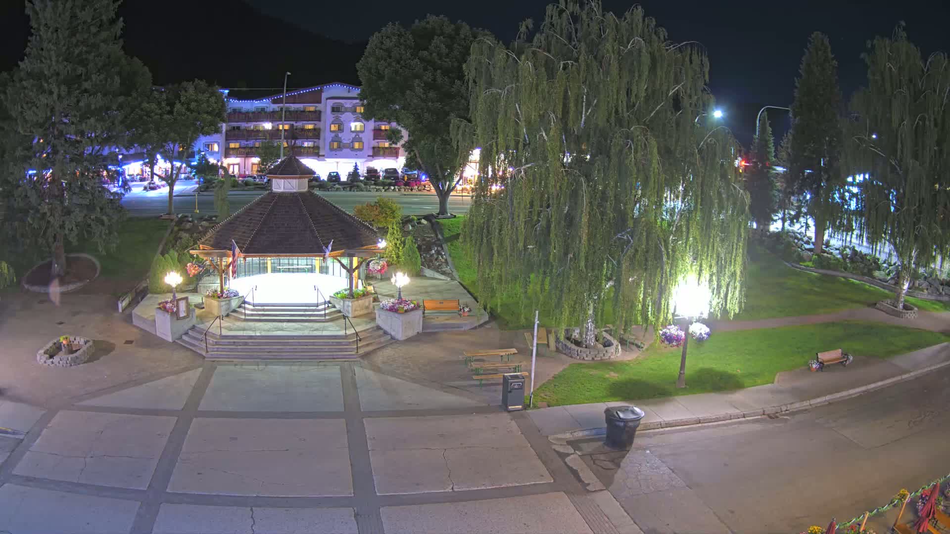 A gazebo sits in a town square at night, illuminated by streetlights, with a hotel and trees visible in the background.