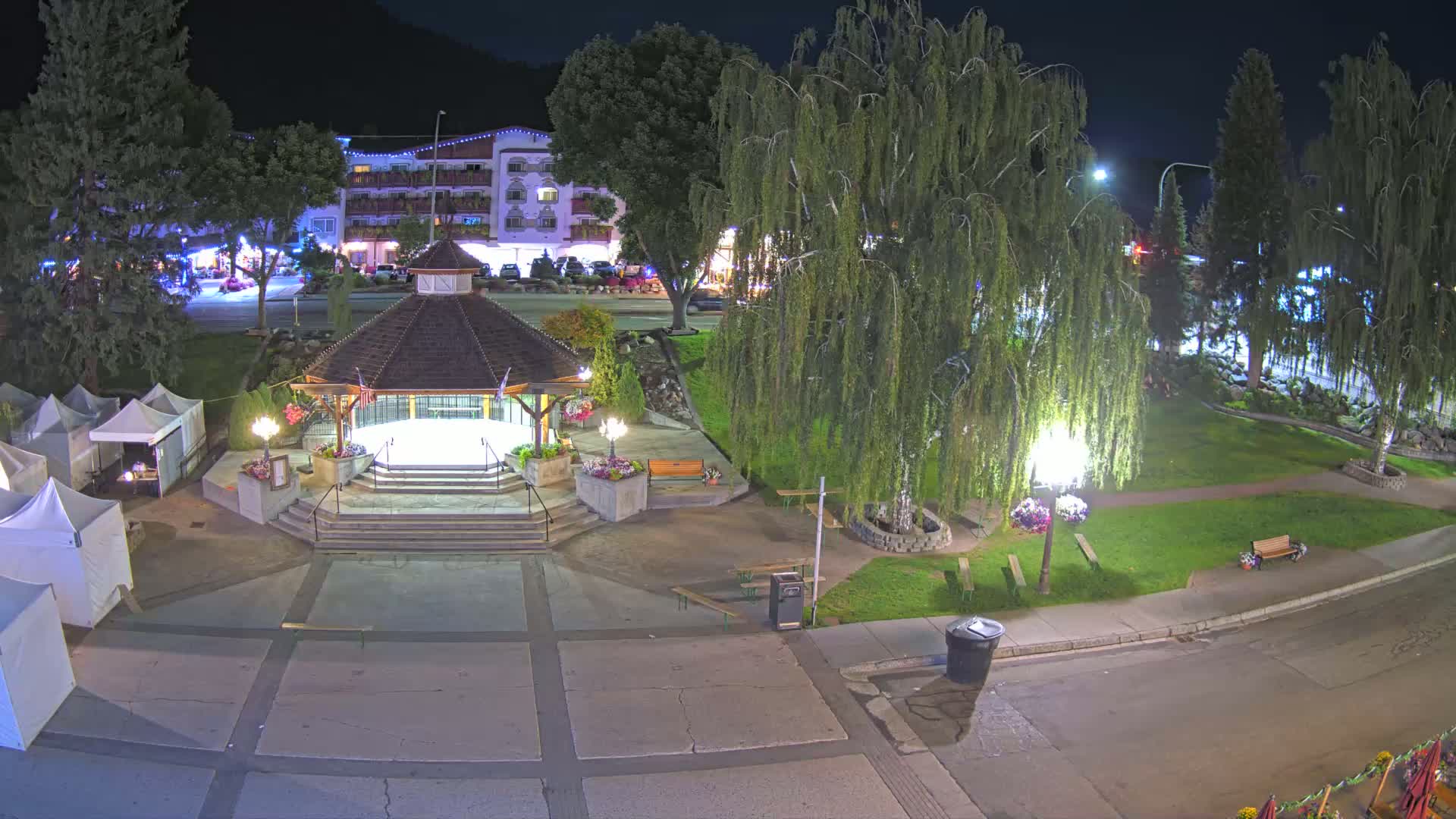 A nighttime view of a town square featuring a gazebo, several trees, some tents, and a building in the background under clear skies.