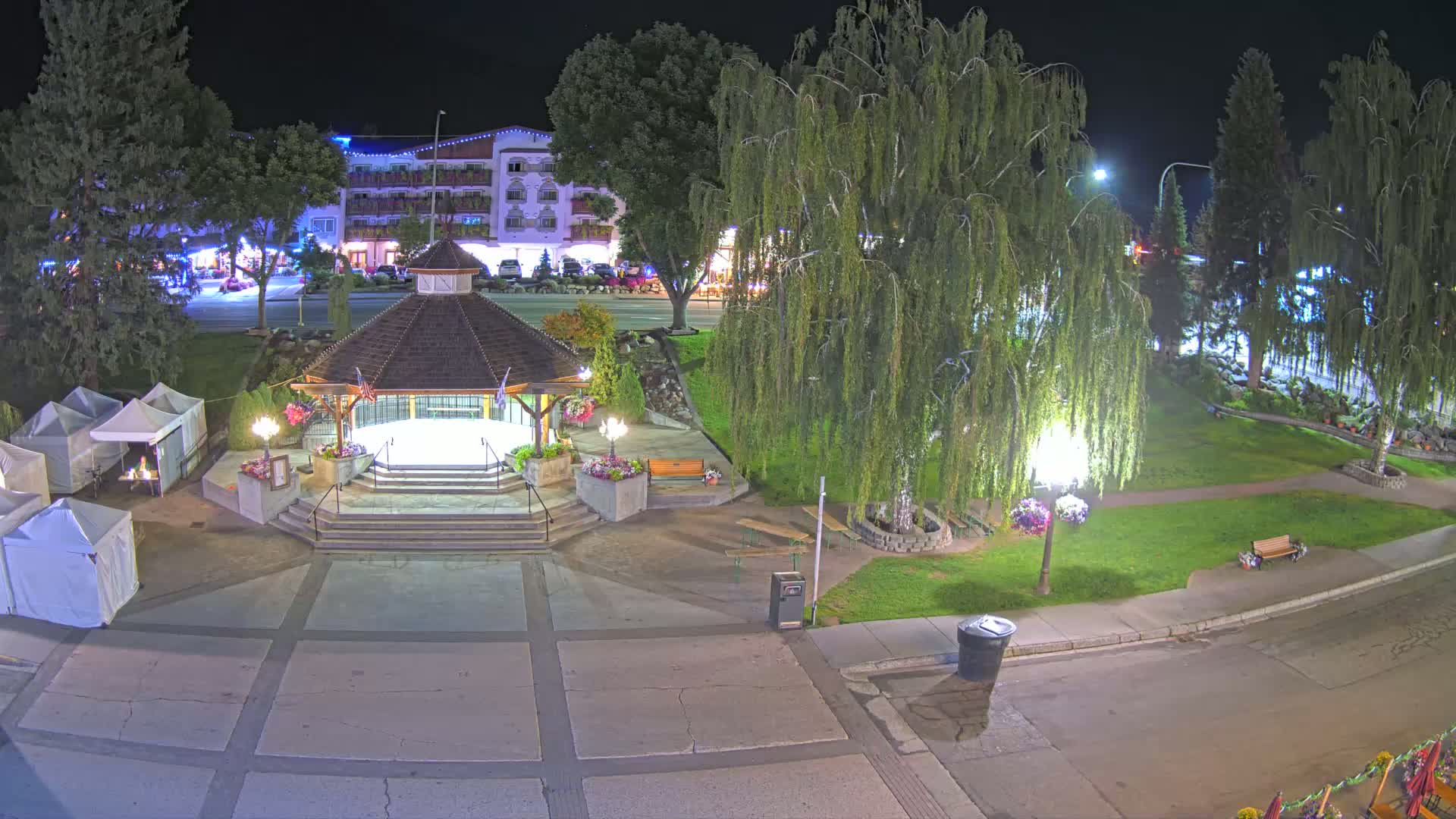 A nighttime view of a town square with a gazebo, several trees, some tents, and a building in the background.