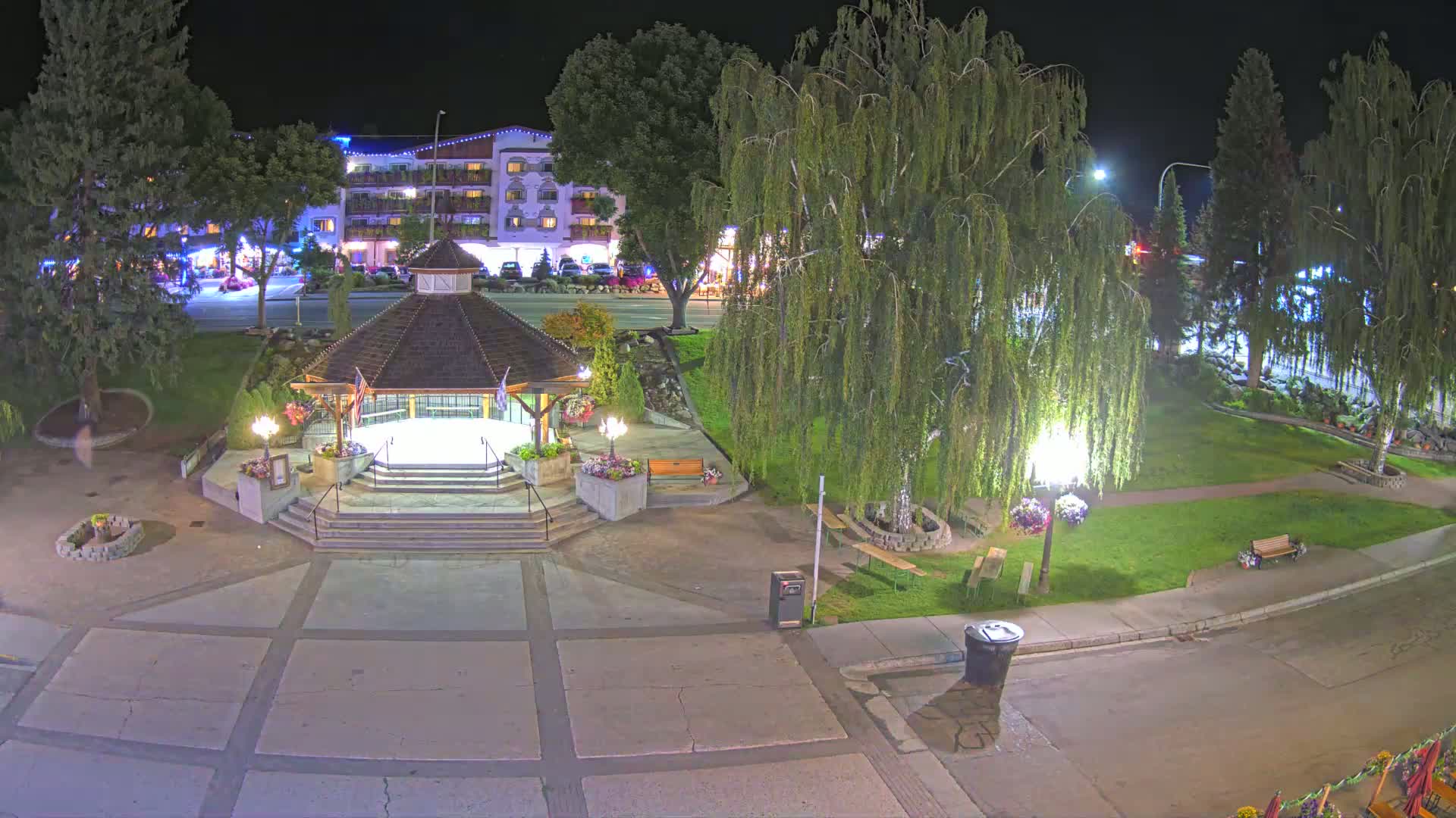 A nighttime view of a town square featuring a gazebo, a weeping willow tree, and a multi-story building in the background.