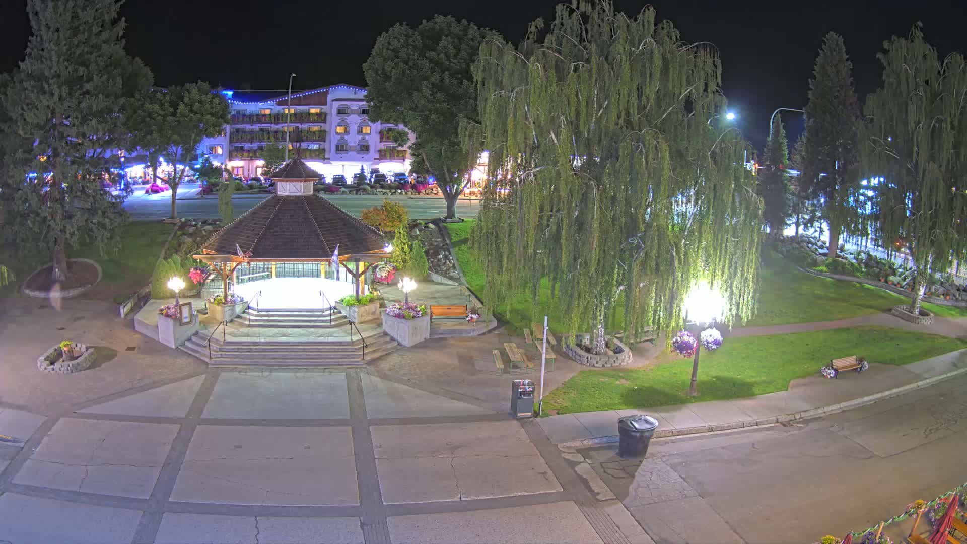 A nighttime, overhead view of a park featuring a gazebo, several trees, and a paved area next to a street with buildings visible in the background.