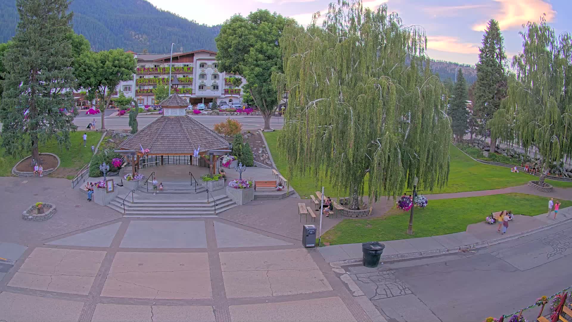 A nighttime, overhead view of a park featuring a gazebo, several trees, and a paved area next to a street with buildings visible in the background.
