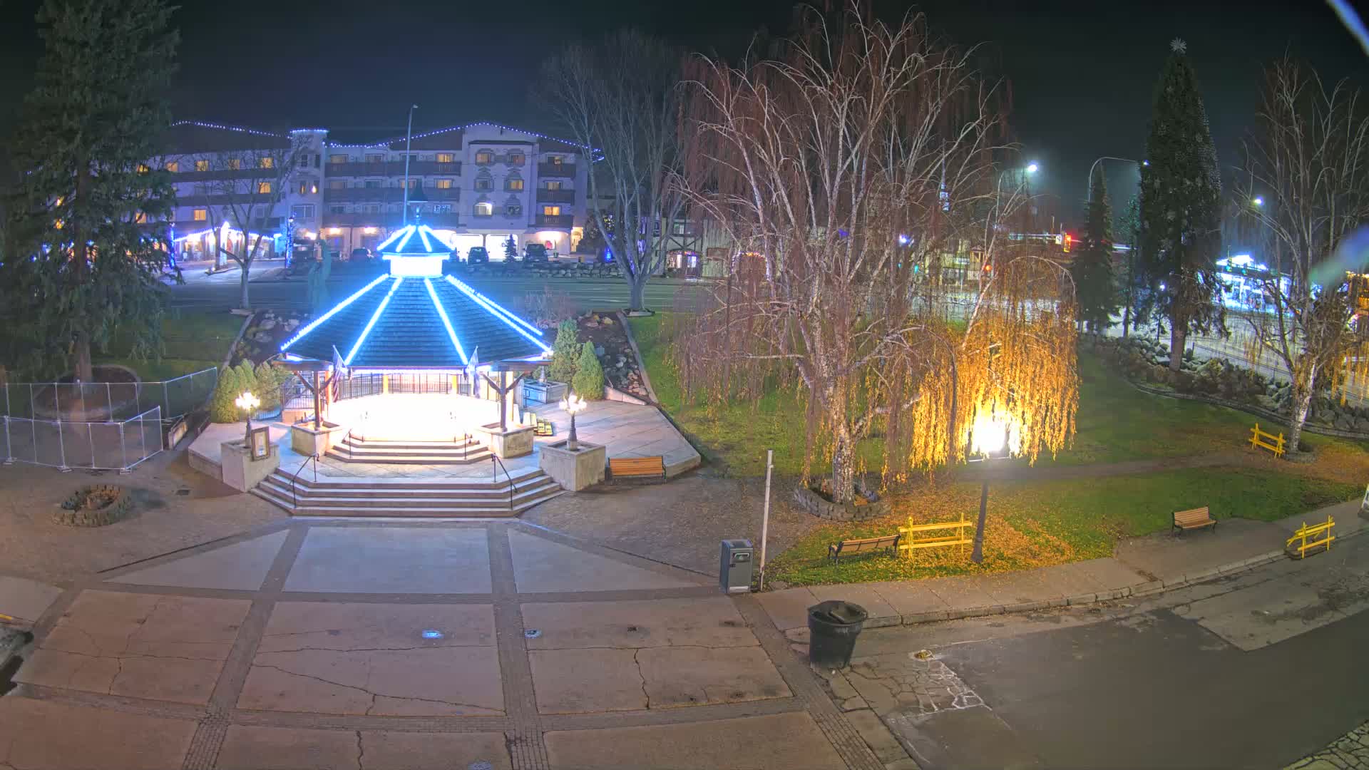 On a clear night, a central gazebo with bright blue lights anchors a paved square, surrounded by grassy park areas with trees—including one illuminated by warm orange lights—and multi-story buildings decorated with white string lights in the background.
