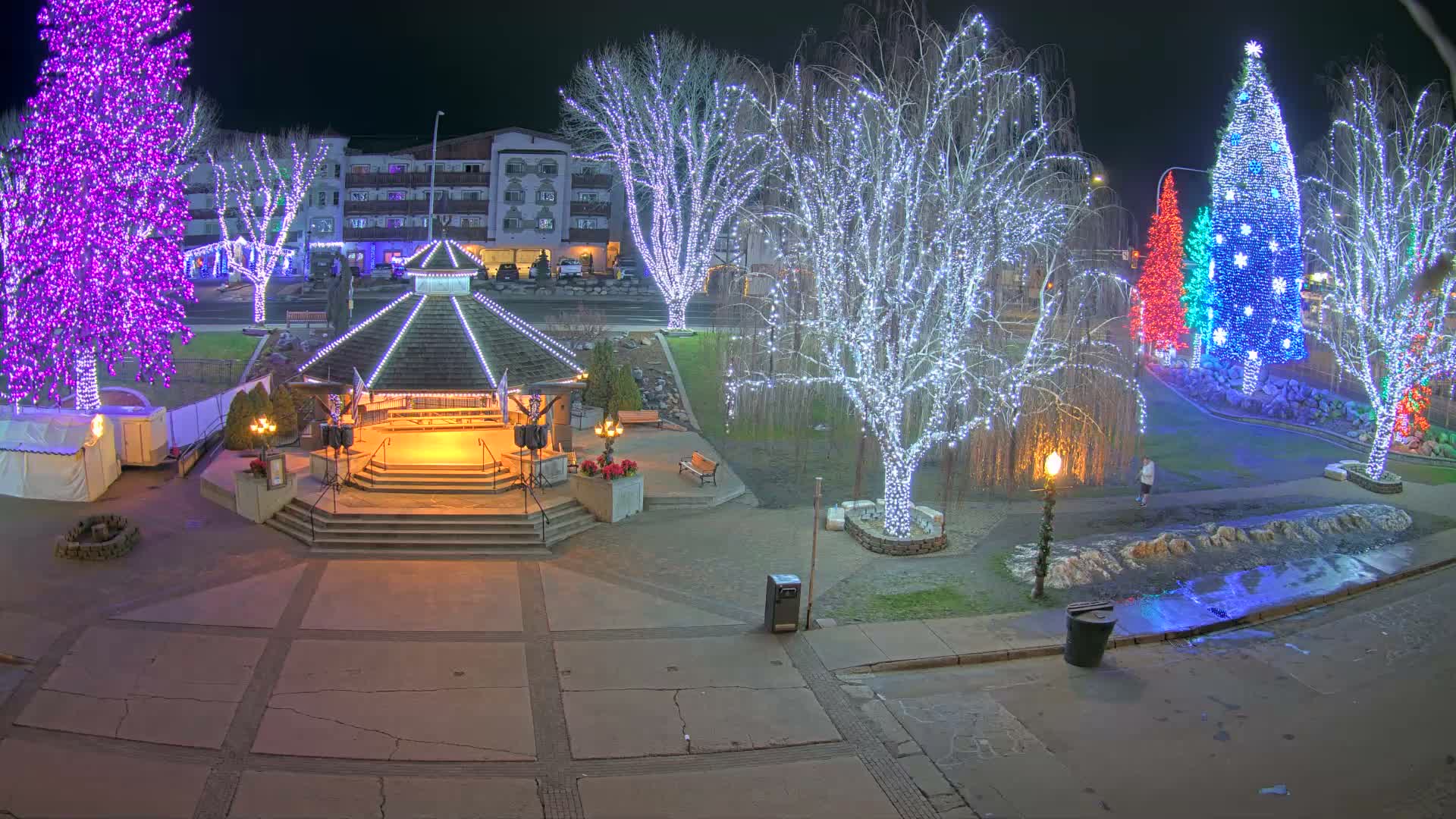 A brightly illuminated town square or park is shown at night, featuring a central gazebo adorned with lights, numerous bare trees wrapped in white and purple string lights, and a distant row of festive trees decorated in red, blue, and green, all under a clear dark sky.