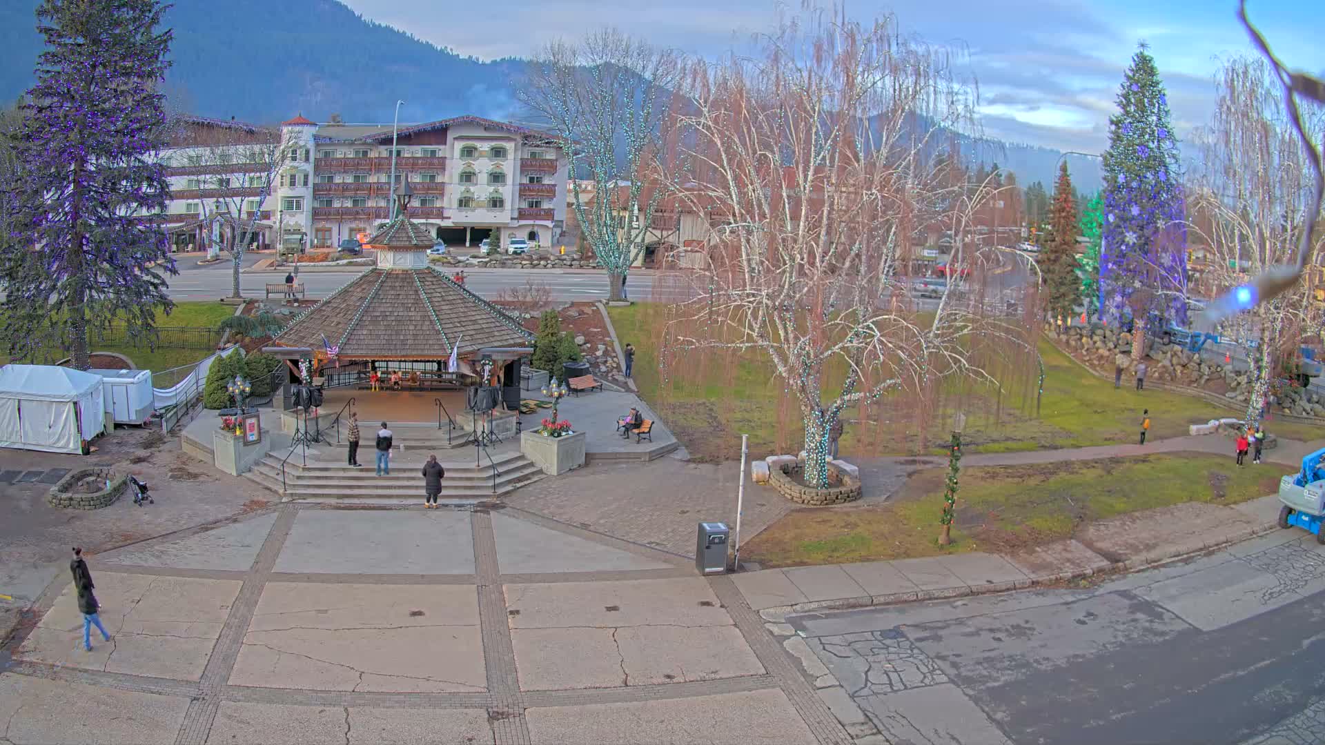 A brightly illuminated town square or park is shown at night, featuring a central gazebo adorned with lights, numerous bare trees wrapped in white and purple string lights, and a distant row of festive trees decorated in red, blue, and green, all under a clear dark sky.