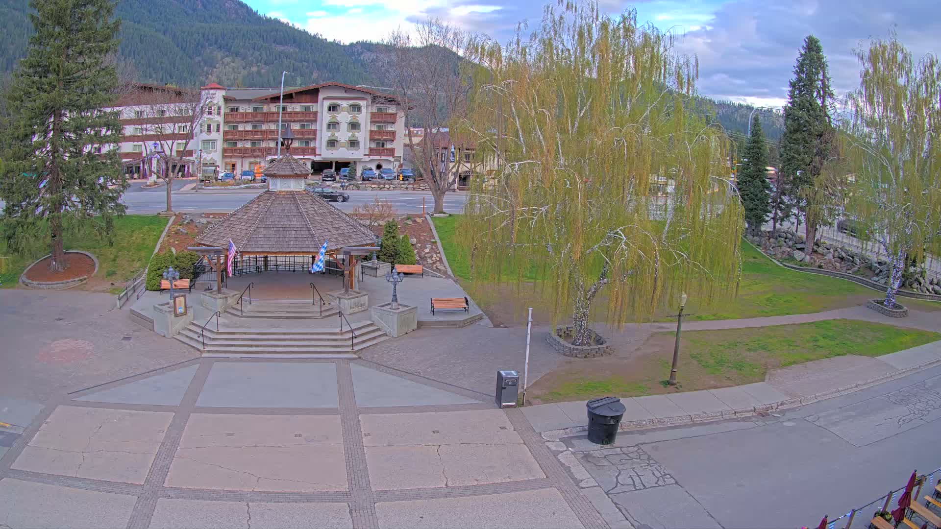 A brightly illuminated town square or park is shown at night, featuring a central gazebo adorned with lights, numerous bare trees wrapped in white and purple string lights, and a distant row of festive trees decorated in red, blue, and green, all under a clear dark sky.