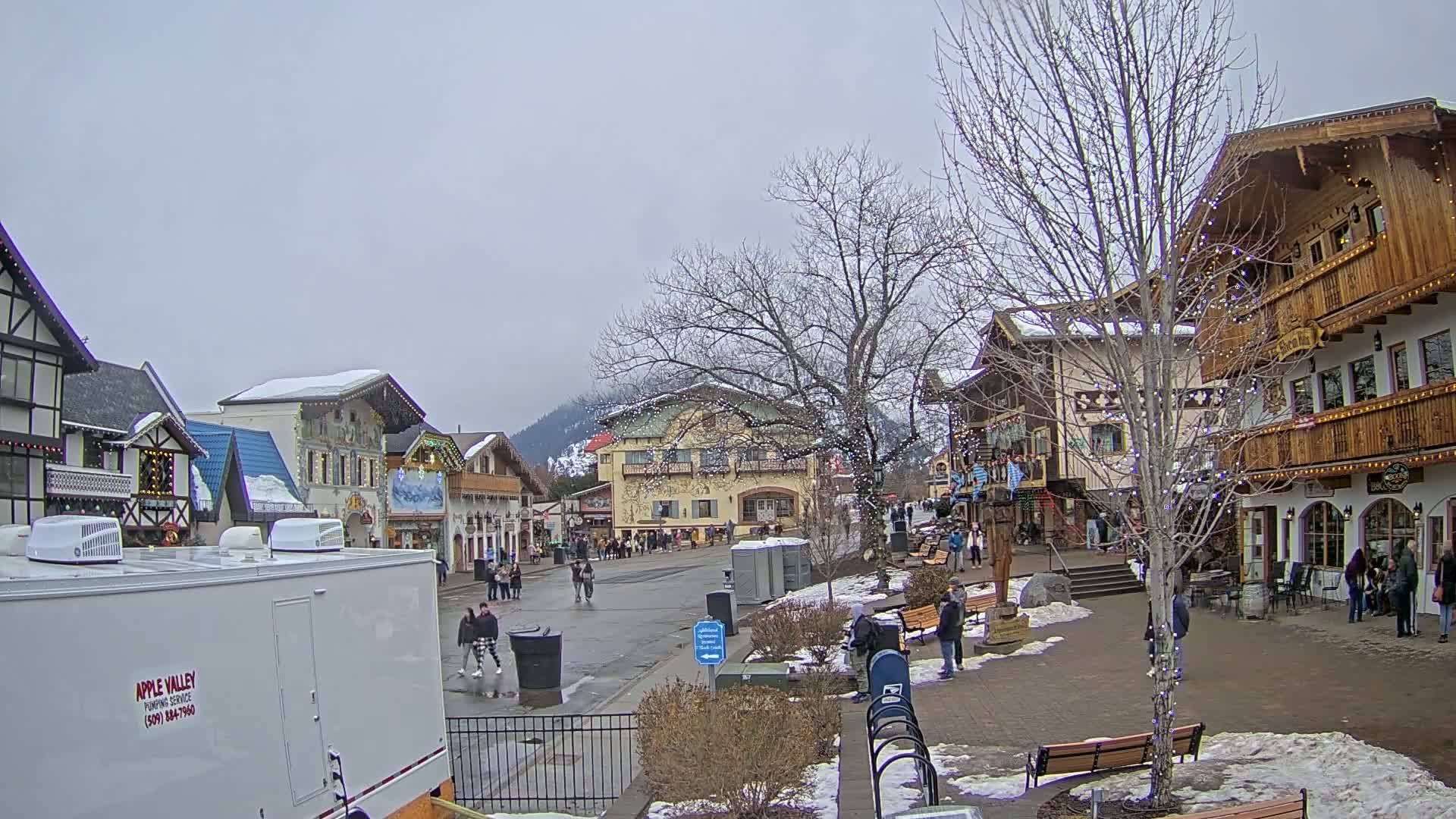 A snow-dusted village street scene shows pedestrians walking past European-style buildings on an overcast day.