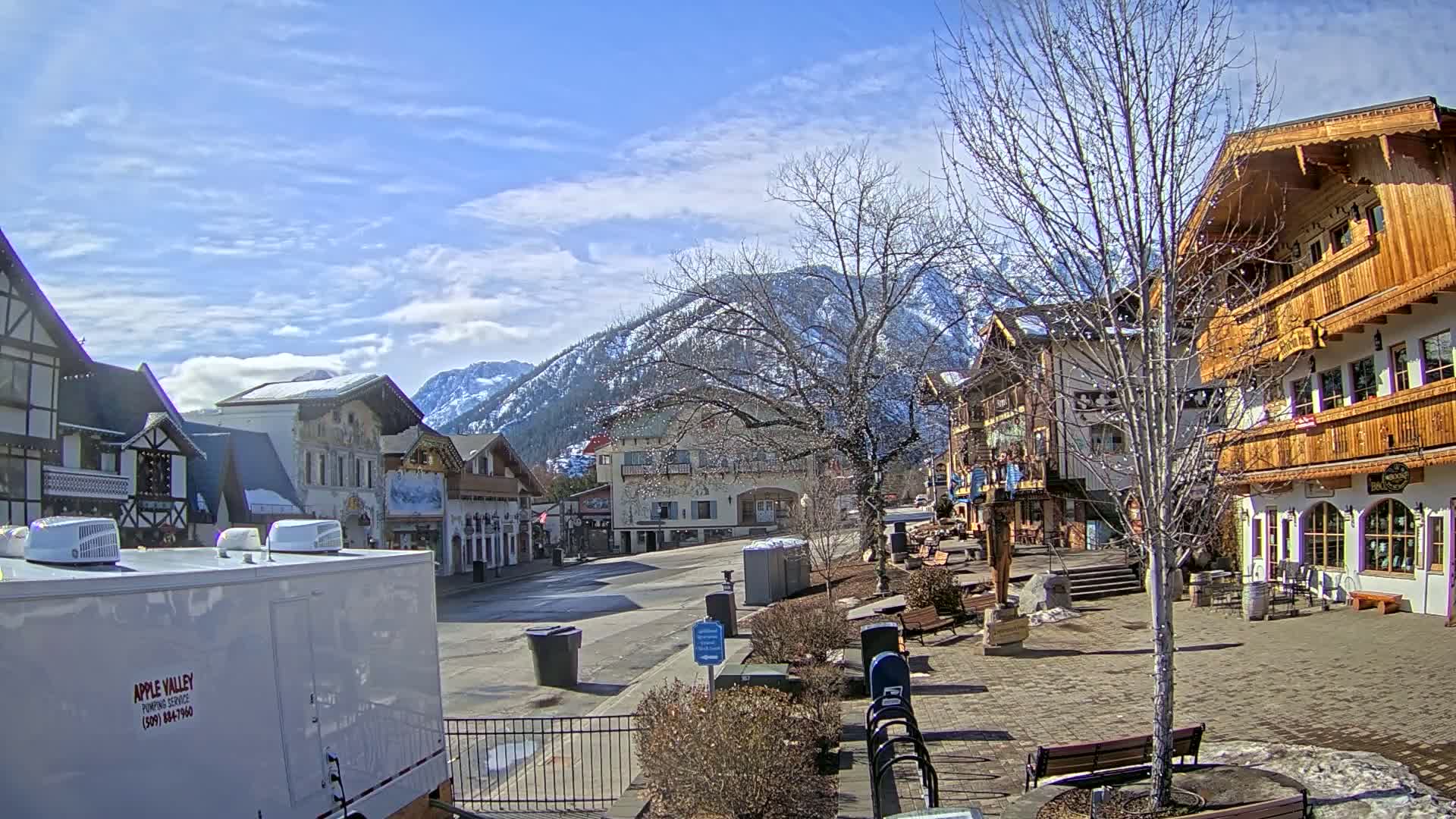 A mostly sunny day reveals a village square with several European-style buildings, a snow-capped mountain in the background, and bare deciduous trees.
