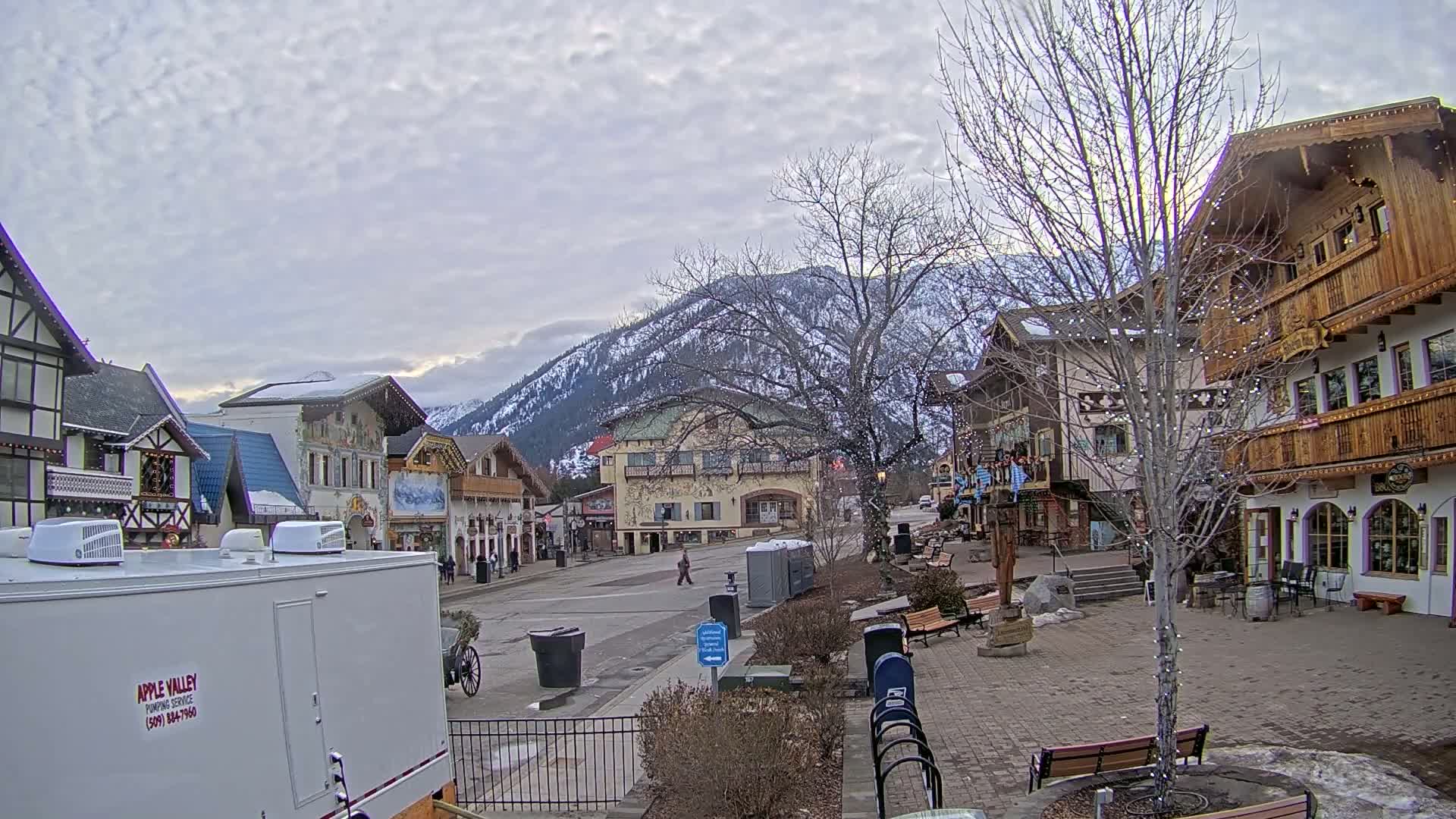 A snow-dusted mountain range overlooks a town square with Bavarian-style buildings under a cloudy sky.