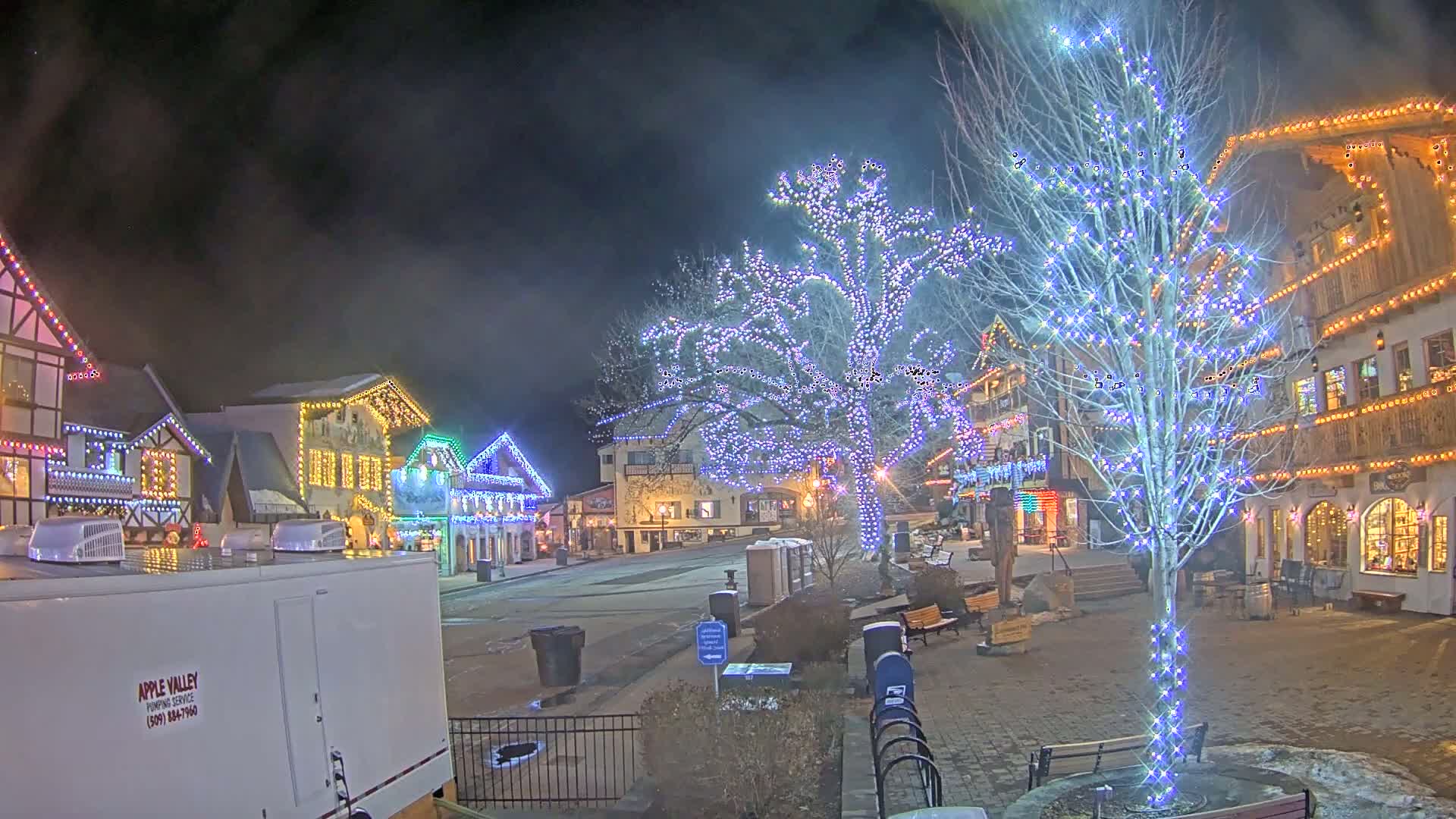 A nighttime scene of a town square decorated with colorful Christmas lights on buildings and trees, under a hazy sky.