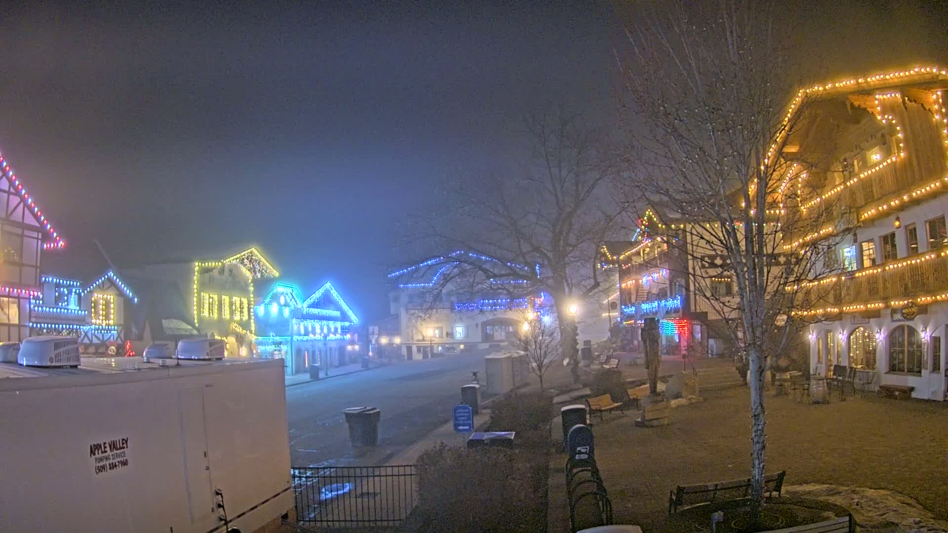 A foggy night scene shows a row of buildings decorated with colorful Christmas lights.