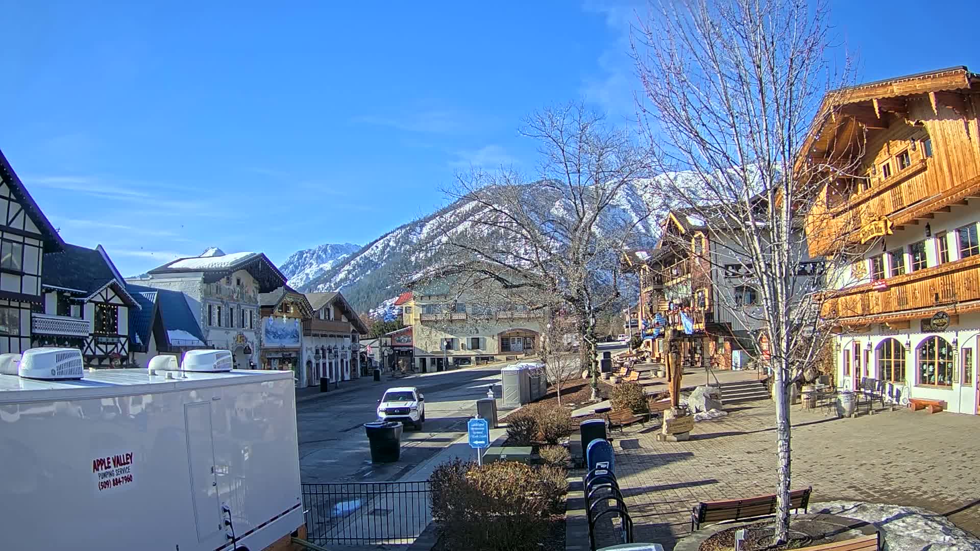 A sunny day reveals a quaint village square nestled against snow-capped mountains, featuring several European-style buildings and a sparse amount of vehicles.