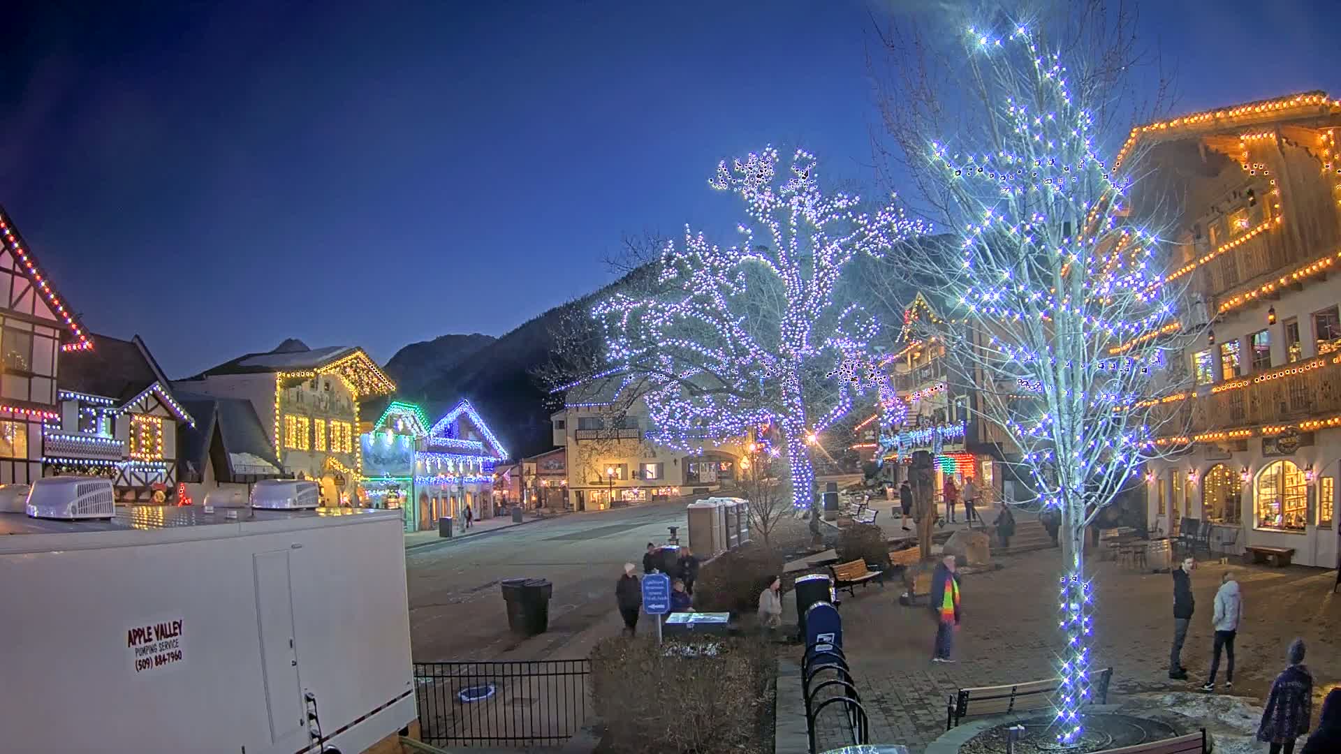 A town square at night, illuminated by colorful Christmas lights strung on buildings and trees, under a clear, dark blue sky.