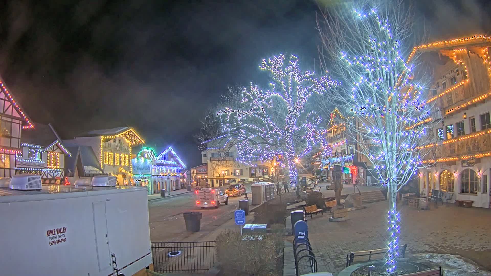 A nighttime scene of a village square adorned with Christmas lights on buildings and trees, with a few cars and people visible.
