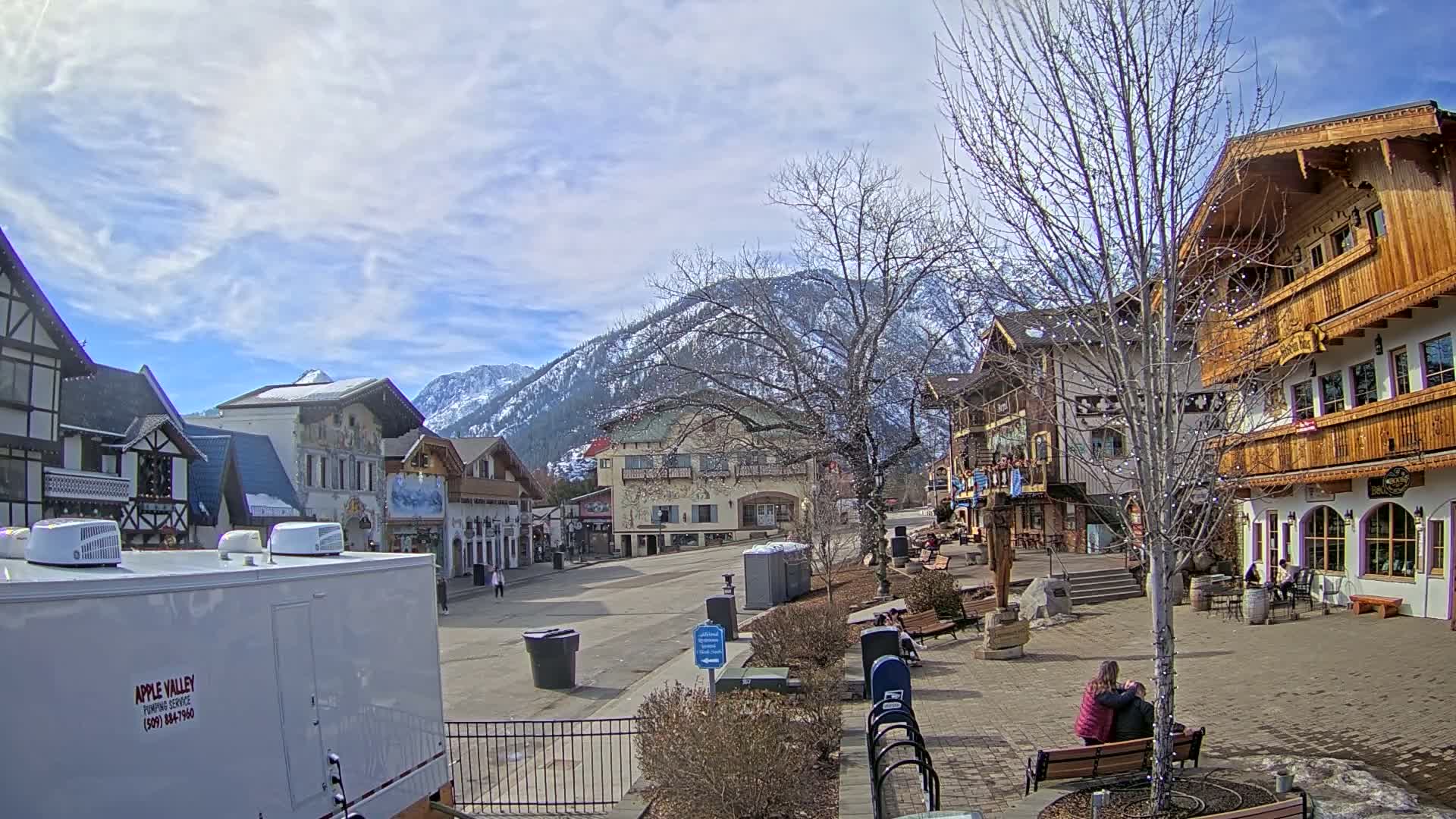 A mostly sunny day with partly cloudy skies shows a quaint village square with several European-style buildings, a snow-capped mountain range in the background, and a few people scattered throughout.