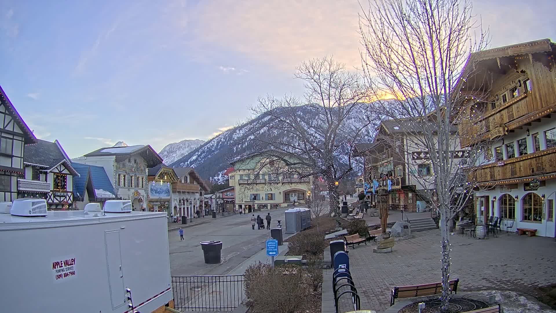 A quaint town square with European-style buildings, under a partly cloudy twilight sky, is nestled against a snow-capped mountain range.