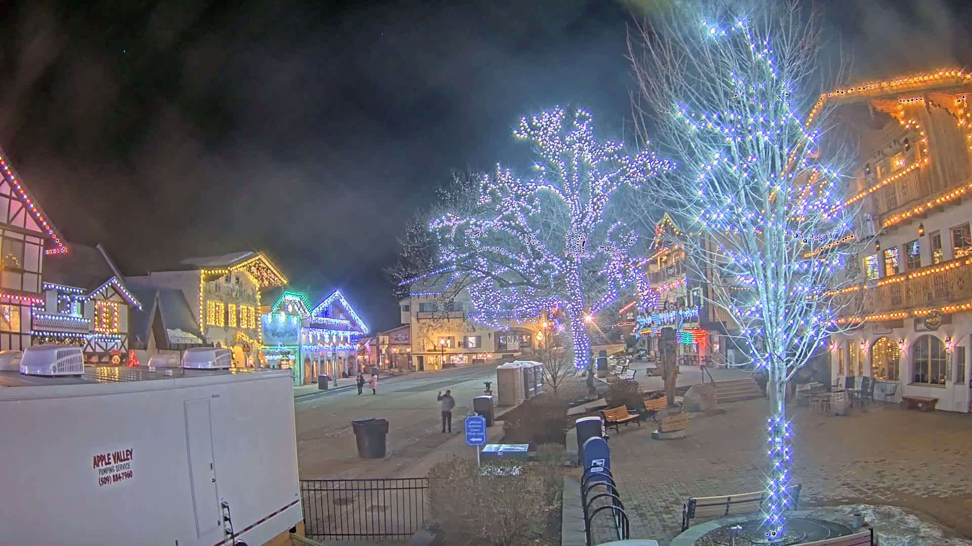 A nighttime scene of a town square decorated with Christmas lights, including illuminated trees and buildings, under a slightly hazy sky.