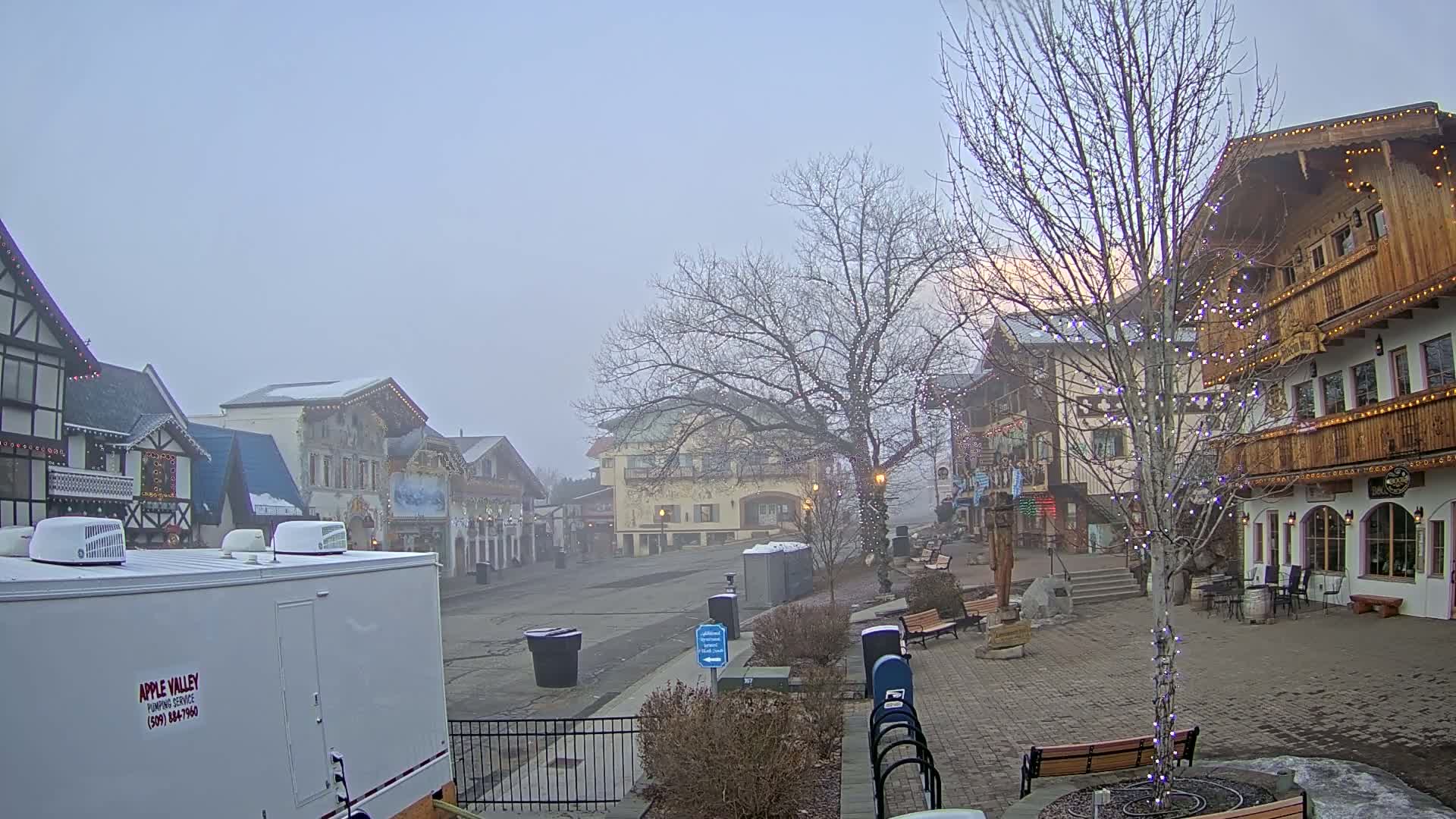 A foggy, snow-covered village square with several multi-story buildings, some decorated with holiday lights, sits mostly empty under a pale sky.