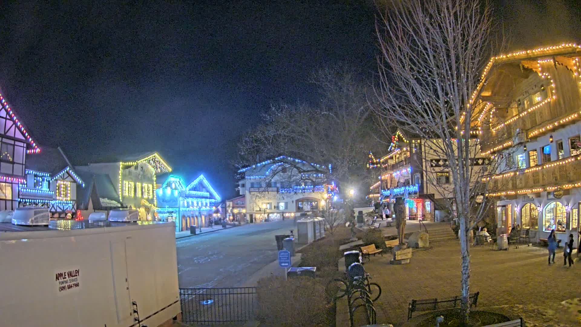 A nighttime scene of a village square with several buildings decorated with colorful lights, some snow on the ground, and bare trees.