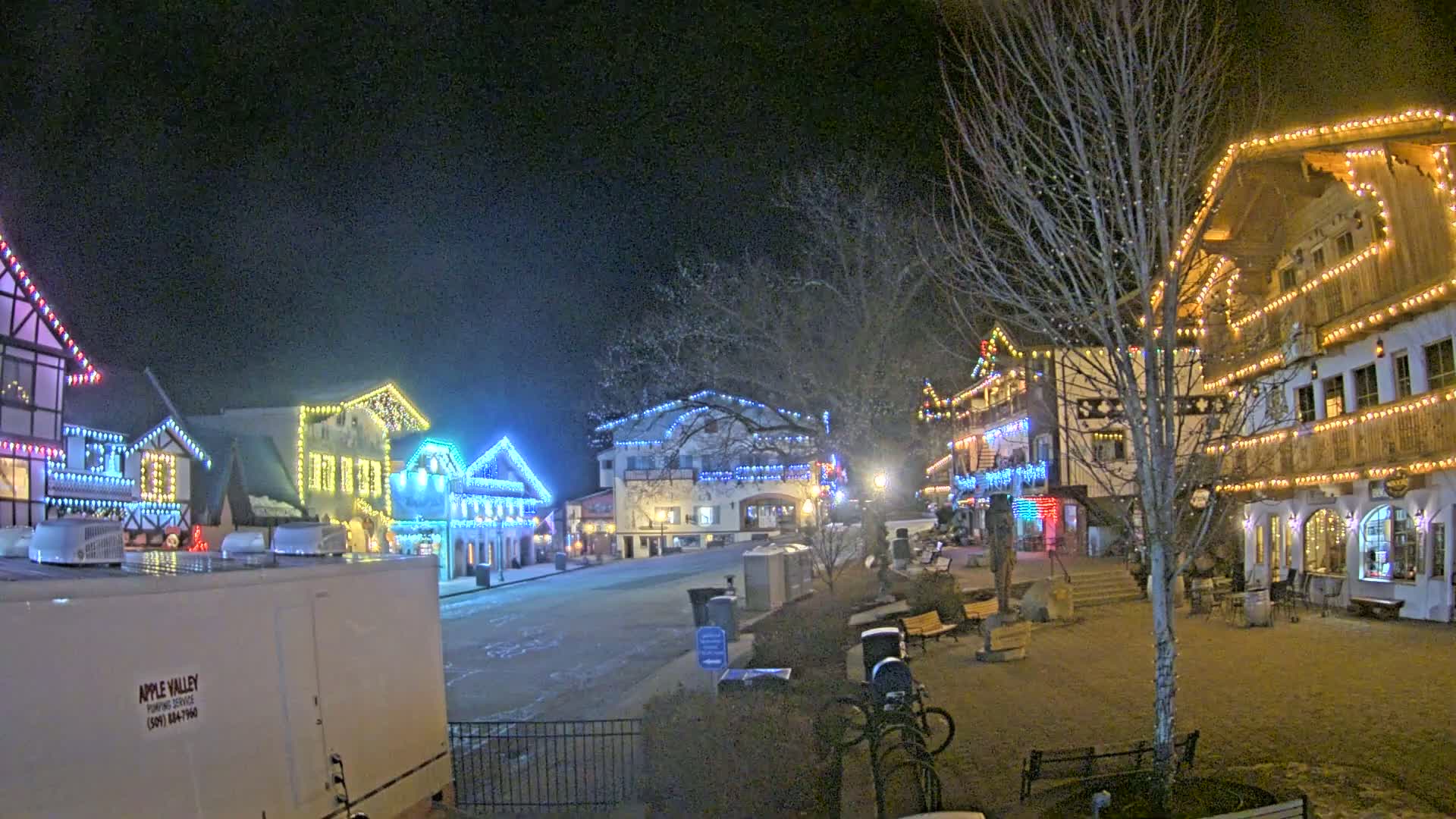 A snow-dusted town square at night, illuminated by colorful lights strung across several buildings.