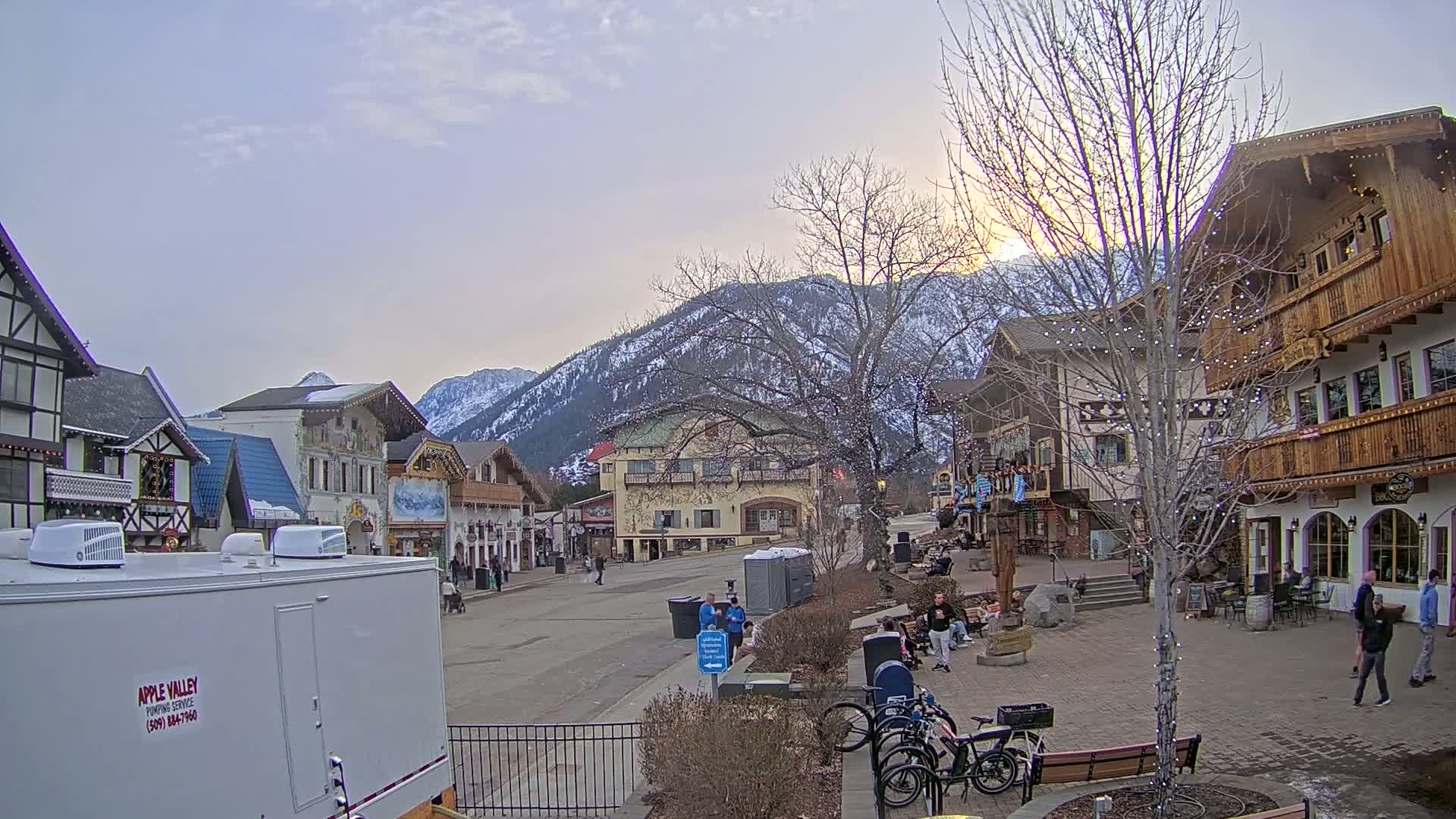 A mostly empty, European-style village street sits beneath a partially cloudy sunset sky, backed by snow-dusted mountains.
