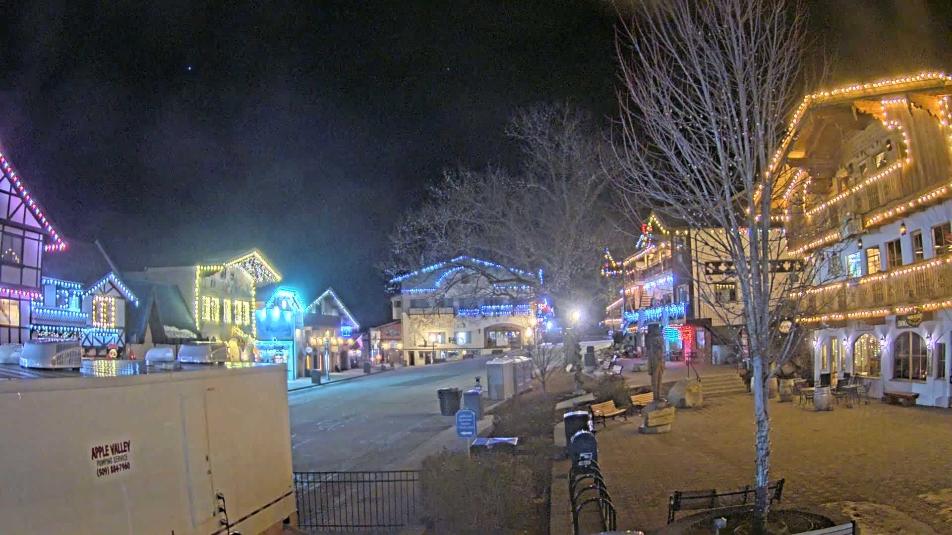 A nighttime scene of a village square with several buildings decorated with colorful lights, under a clear, dark sky.