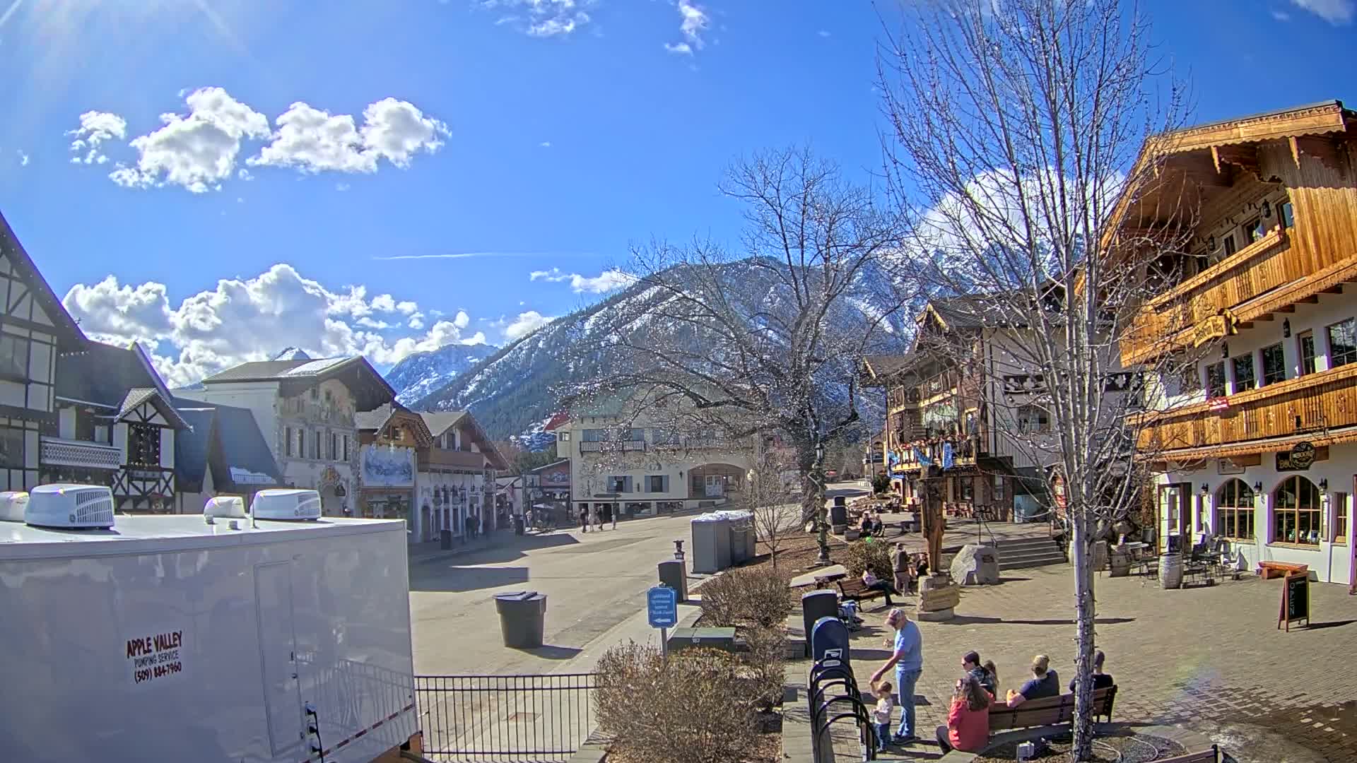 A sunny day in a Bavarian-style village, nestled against snow-capped mountains, shows several people relaxing outdoors.