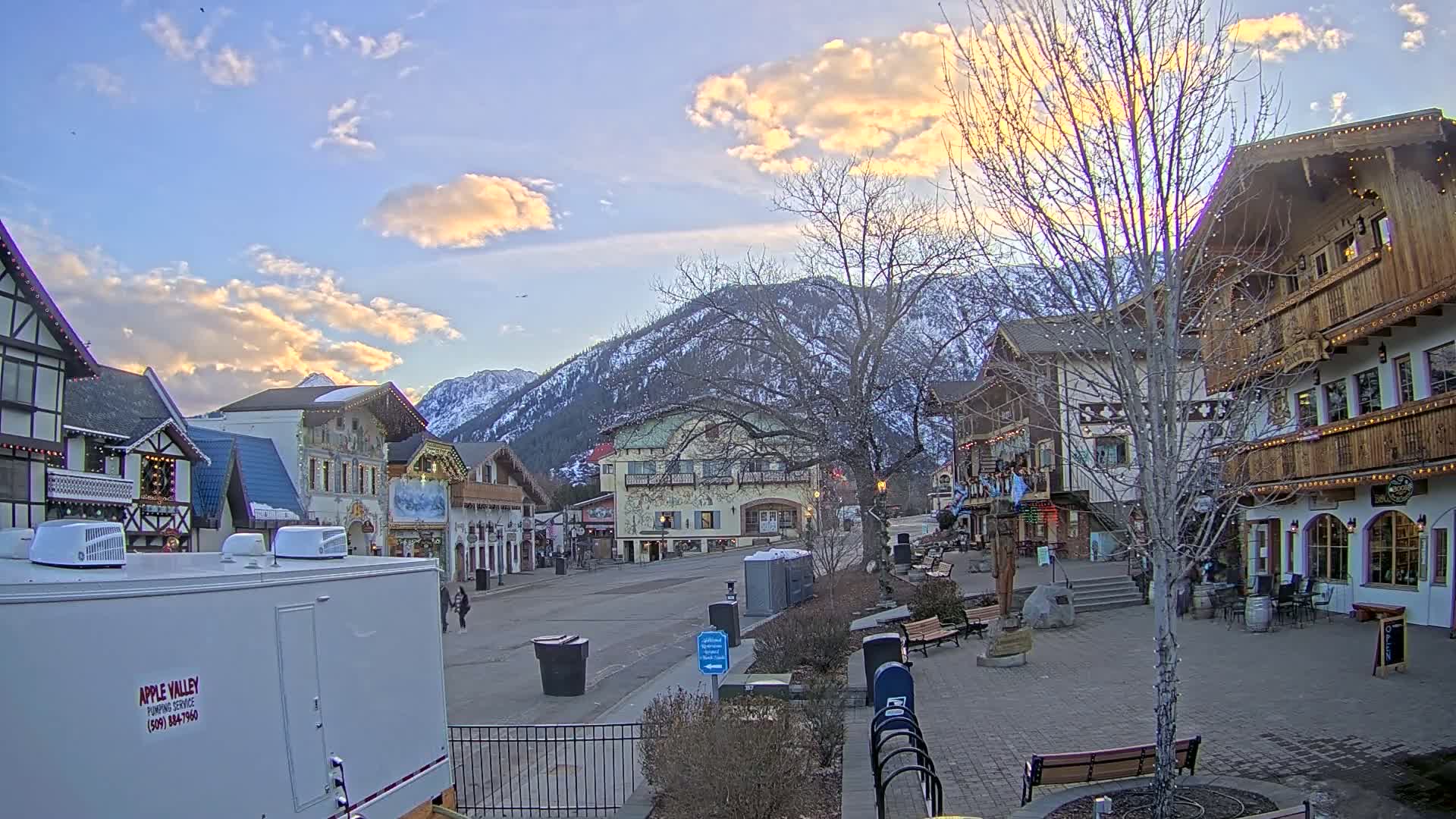 A quaint, European-style village street scene at sunset, nestled against snow-capped mountains under a partly cloudy sky.