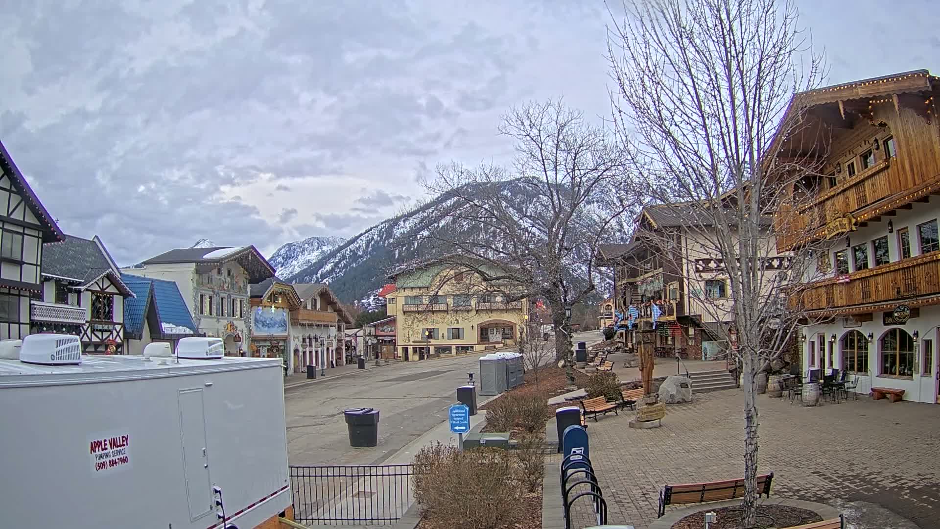 A mostly empty town square with European-style buildings and snow-capped mountains in the background under a cloudy sky.