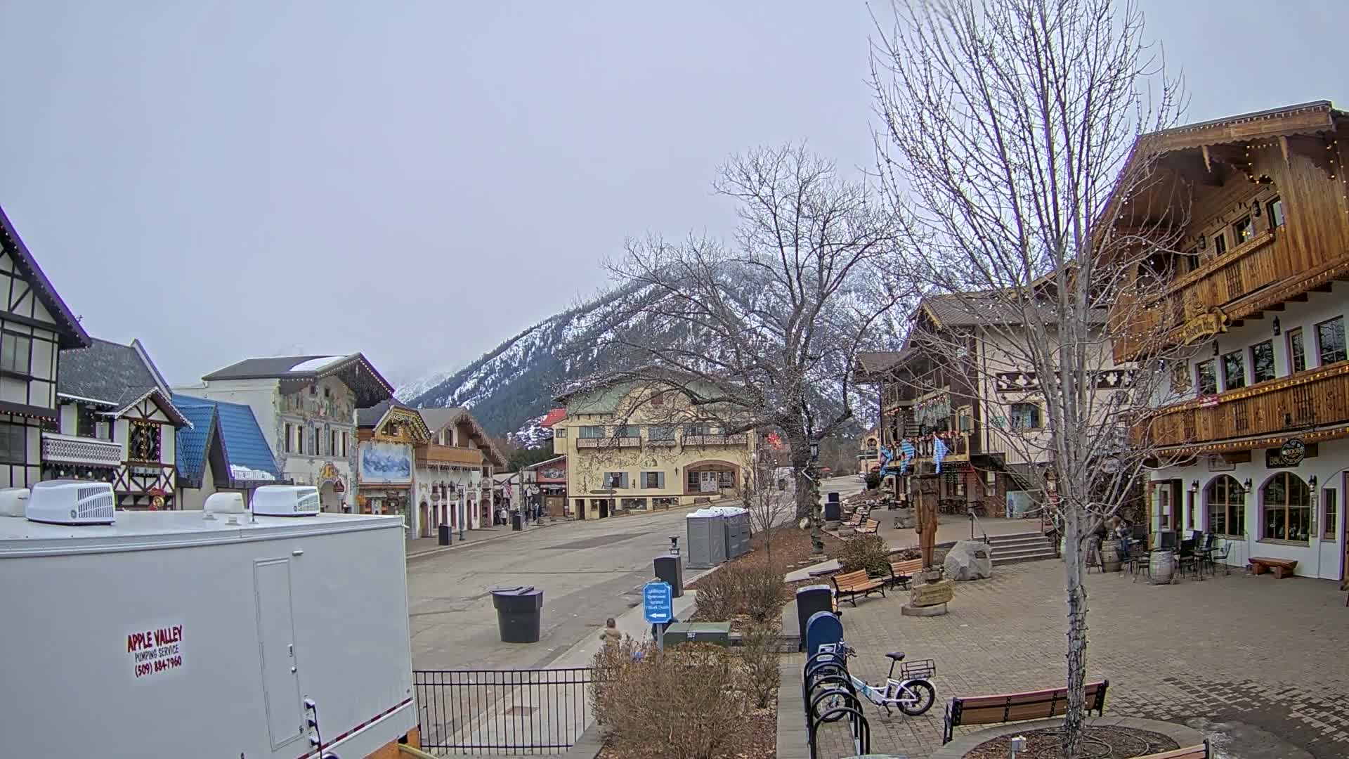 A mostly empty town square with snow-dusted mountains in the background under an overcast sky, features several Bavarian-style buildings and a few parked bicycles.