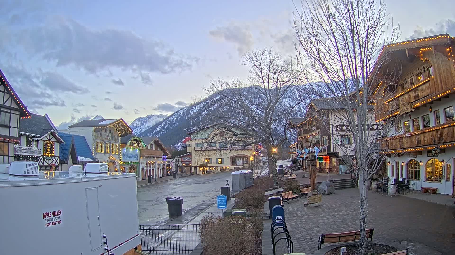 A mostly empty town square with European-style buildings decorated with lights, under a partly cloudy twilight sky, is situated against a backdrop of snow-capped mountains.